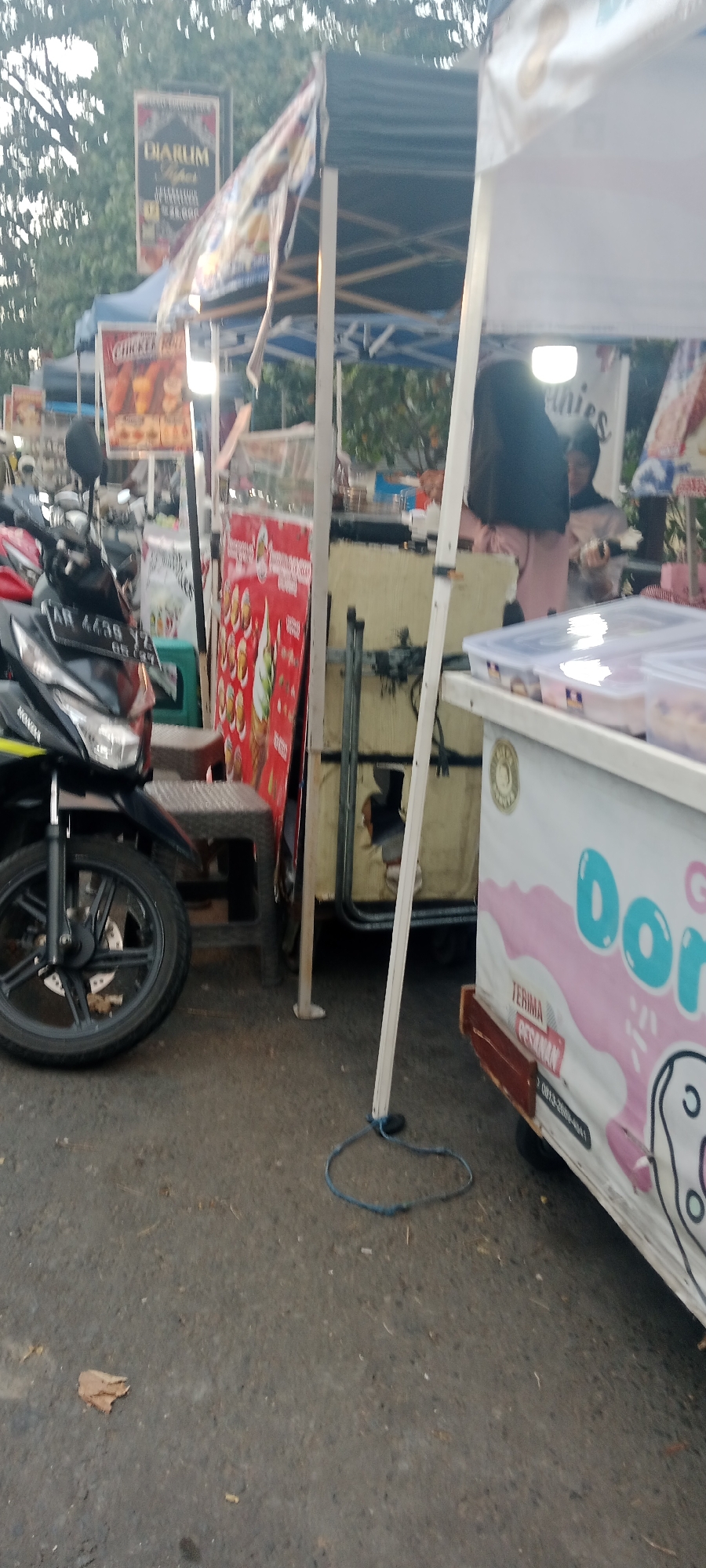 This image depicts an outdoor street scene in Ngemplak, Indonesia, likely a market or roadside vending area. In the foreground, a motorcycle is parked on the asphalt ground. Behind the motorcycle, a food stall is set up with various items. A red sign displaying images of ice cream cones is prominently displayed. Further back, another stall features a person, partially obscured, attending to food. A white tent canopy provides shade over some of the stalls. The overall environment appears to be a busy commercial street with trees in the background.