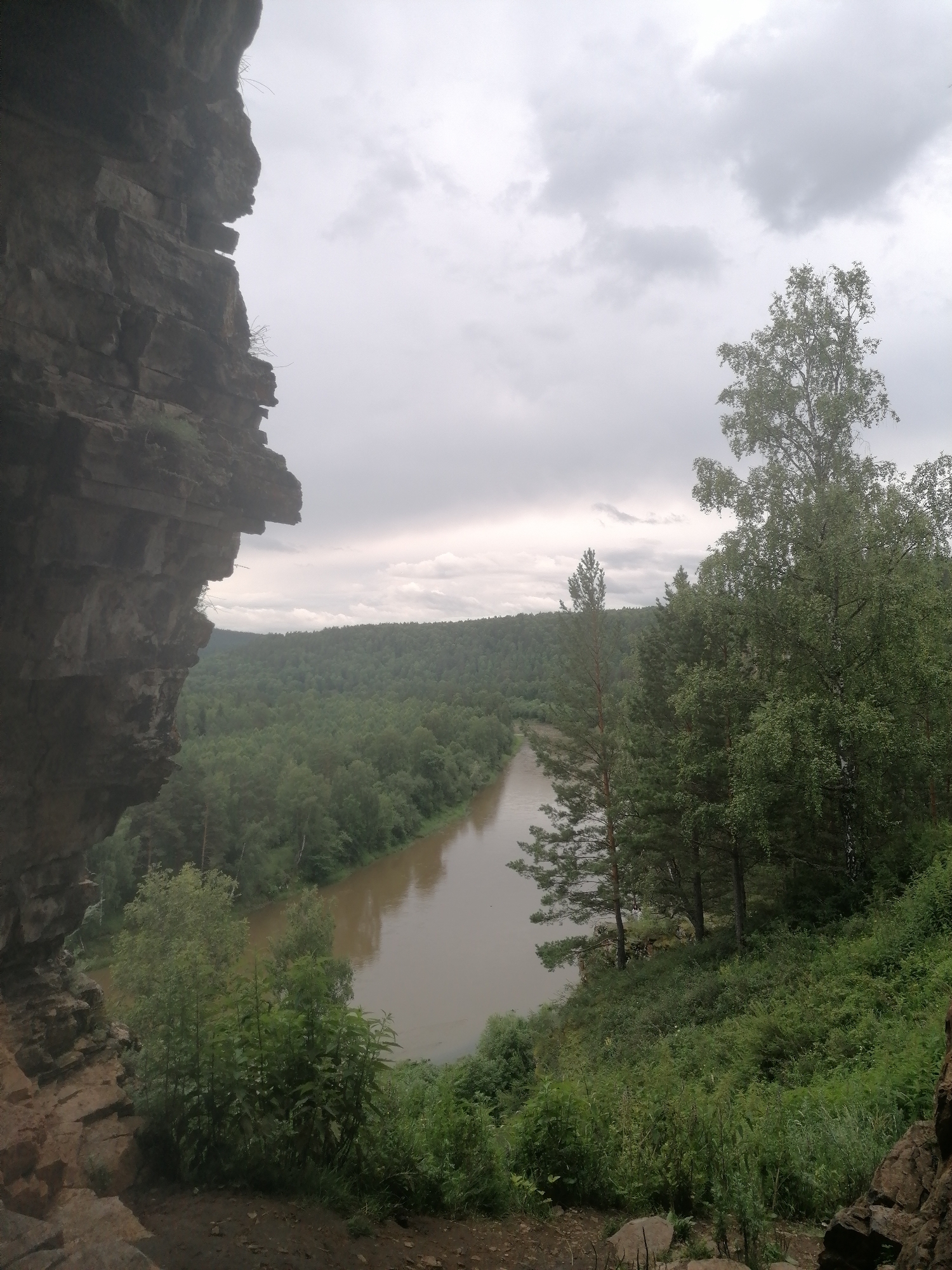 This media file depicts a serene natural landscape from a high vantage point, looking out over a wide river valley. On the left, a towering, weathered rock face dominates the foreground, its rough texture and stratified layers suggesting a long geological history. The rock face creates a sense of depth and perspective, framing the view beyond.
The valley floor is blanketed in a dense, verdant forest that stretches as far as the eye can see, with a multitude of trees creating a textured, green expanse. A calm, wide river meanders through the center of the valley, its surface reflecting the overcast sky. The water appears muddy or silty, suggesting it might be a slow-moving river or one carrying sediment.
On the right side of the frame, lush green foliage and trees grow on a slope leading down to the river. The trees are a mix of deciduous and coniferous types, with some taller specimens standing out against the more uniform forest canopy.
The sky is filled with heavy, grey clouds, indicating an overcast or perhaps a looming storm. This diffused light contributes to a subdued and peaceful atmosphere. The time of day is indeterminate due to the cloudy conditions, but it appears to be daytime. The overall mood is tranquil and somewhat wild, highlighting the untouched beauty of the natural environment. There are no visible people or objects other than the natural elements. The location is identified as Lagerevo, Russia.