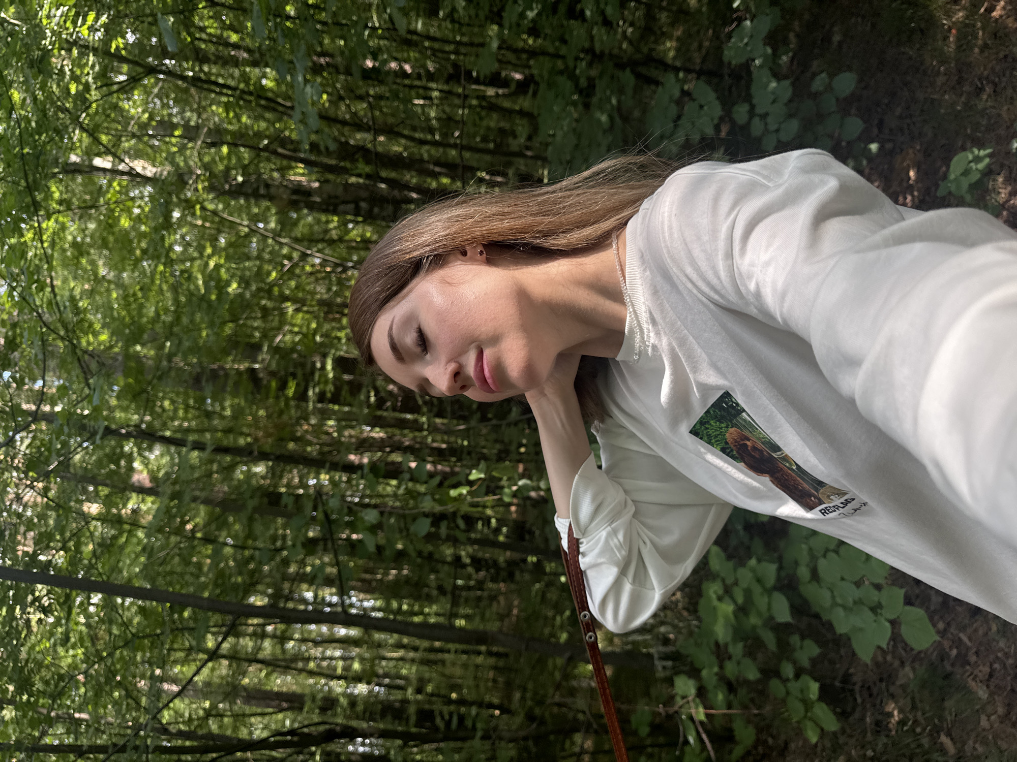 A young woman with shoulder-length blonde hair is standing in a forest, with her eyes closed and her head tilted back, as if enjoying the sun. She is wearing a white long-sleeved shirt with a picture of a dog on it and a thin beaded necklace. Her right hand is raised to her neck, and she is holding a thin brown leash with metal grommets in her left hand. The forest is dense with tall trees and green foliage, suggesting a natural, outdoor setting. The lighting indicates it is daytime, with sunlight filtering through the trees. The environment appears to be a typical forest found in the city of Chaikovsky, Russia, given the presence of birch and pine trees, common to the region.