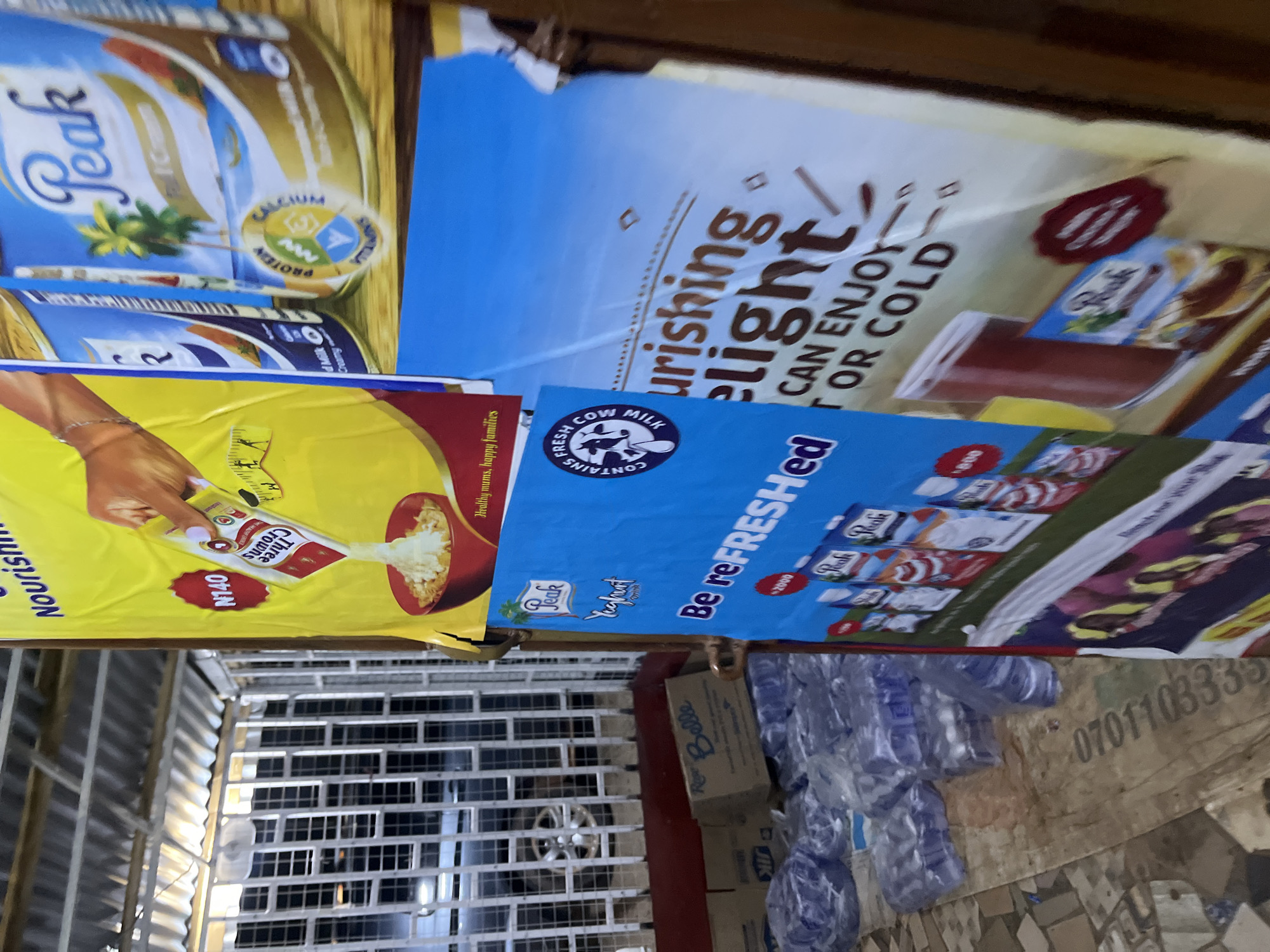 The image depicts an outdoor stall in Bukuru, Nigeria, showcasing various food and beverage advertisements. The stall is covered by a corrugated metal roof, and the background features a white metal grate with a car visible through it.
Dominating the scene are several large posters. A prominent yellow poster advertises "Three Crowns" milk, highlighting its price of N140 and showing milk being poured into a bowl of cereal. Beside it, a blue poster for "Peak" milk features illustrations of palm trees and text mentioning "PROTEIN CALCIUM VITAMINS." Another blue poster, positioned lower, advertises "Peak Yoghurt" with the tagline "Be reFRESHed" and depicts several cartons of the product. Further down, a partially visible poster shows a glass of chocolate drink and a smaller packet of "Peak" milk.
In the foreground, stacks of bottled water in plastic wrapping are arranged on the ground, suggesting they are for sale. A cardboard box labeled "Rev Belle" is also visible amongst the water bottles. The ground is a mix of tiled and unpaved surfaces, with a piece of brown tarp or sheeting bearing a serial number like "070110333" visible on the left side of the image. The overall scene suggests a retail or roadside vending point.