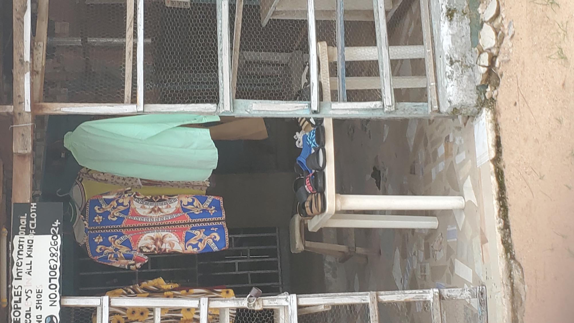 The image shows an outdoor clothing and shoe stall in Jalingo, Nigeria. A sign above the stall reads "PEOPLES INTERNATIONAL" and "DEALERS IN ALL KIND OF CLOTH AND SHOES." A phone number, "NO.07062826024", is also visible on the sign. Several items of clothing are hanging on display, including a brightly colored dress with a baroque pattern and a plain mint green shirt. A white plastic table in front of the stall displays an assortment of sandals and flip-flops. The ground is paved with mosaic tiles, and a wooden stool is placed behind the table. The stall appears to be located on a street, with a rough, sandy area visible in the foreground. The structures around the stall are made of wood and wire mesh.