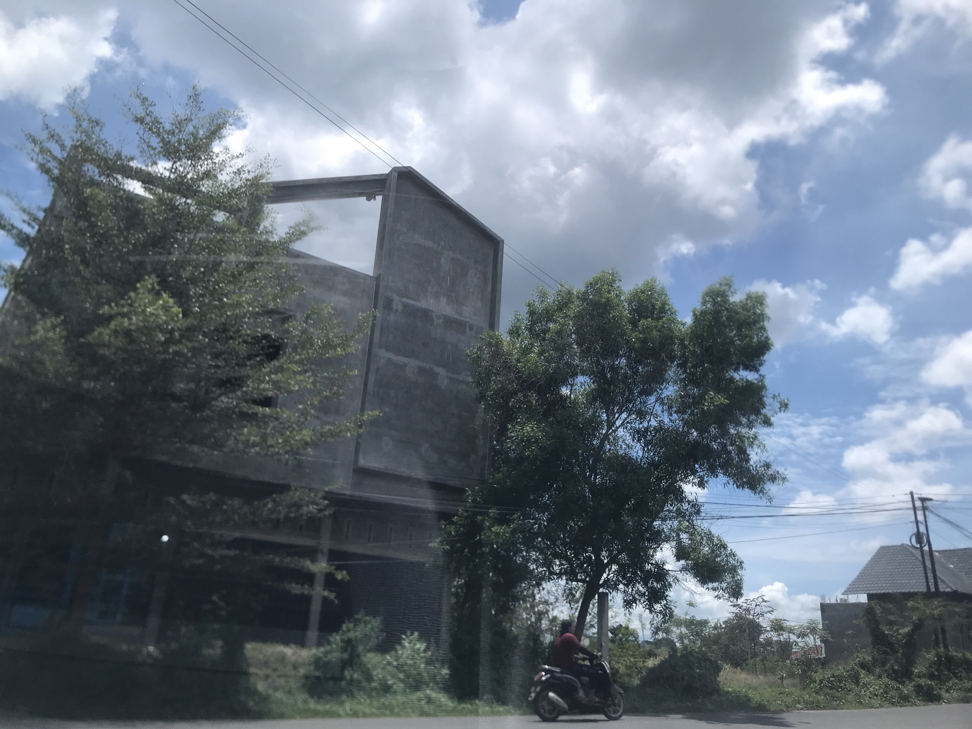 A medium, eye-level shot taken from inside a vehicle on a road reveals a partially constructed, unfinished grey concrete building adjacent to a tree obscuring part of the facade. To the right of the building, a tree and lush greenery surround a motorcyclist. The rider is wearing a dark shirt or jacket. Further in the background is a house with a dark roof, partially obscured by vegetation, suggesting the outskirts or a developing area of Lhokseumawe, Indonesia. Overhead, power lines stretch across a sky filled with scattered clouds and patches of blue sky. A sense of motion is captured, likely from the moving vehicle.