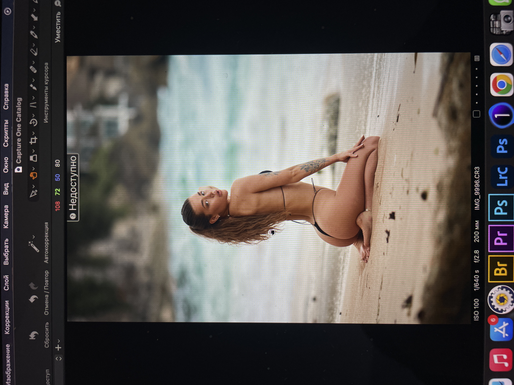 The image shows a photograph of a woman in a black bikini kneeling on a sandy beach. The background features blurry buildings and foliage, suggesting a coastal or lakeside setting. The photo appears to be displayed on a computer screen, showing the interface of a photo editing software. The exposure settings (ISO 100, 1/640 s, f/2.8, 200mm) are visible in the lower right corner, along with icons for programs like Adobe Photoshop, Premiere Pro, and Lightroom. The image is tilted, and the woman is facing the viewer. There's a tattoo on her left arm. The city of Kazan, Russia, is not evident in the image.
