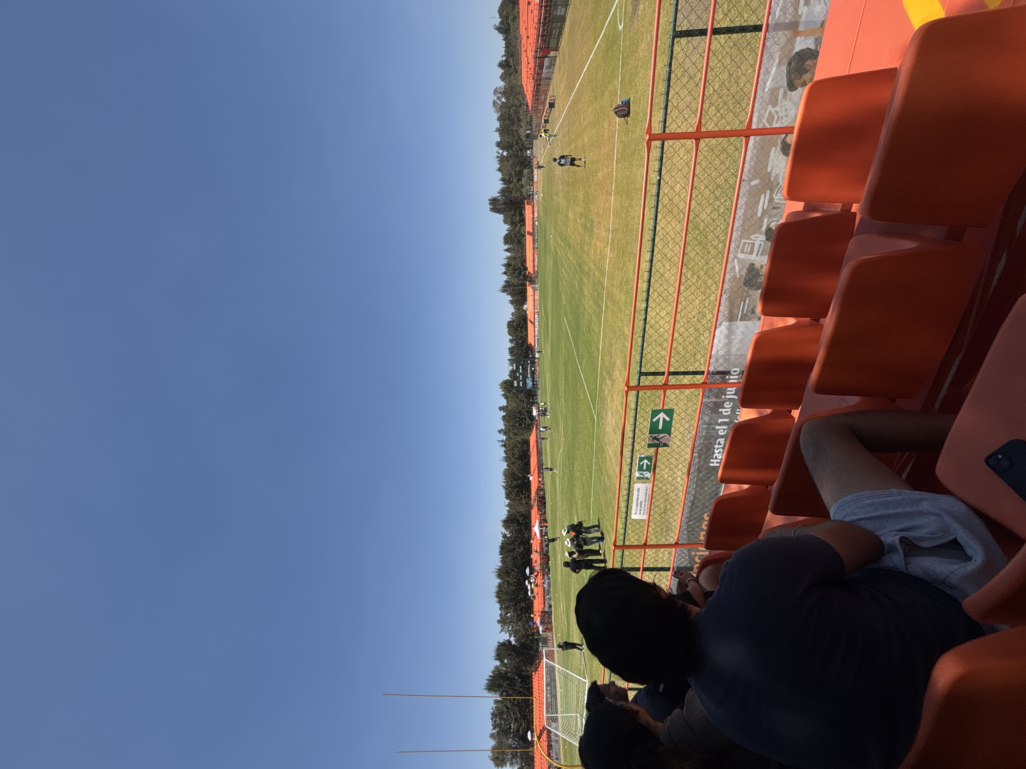 The image captures an outdoor sports stadium under a clear blue sky in San Andrés Cholula, Mexico, likely part of the Universidad de las Américas Puebla (UDLAP) campus, as indicated by a partial sign. The scene is viewed from behind three spectators seated in orange stadium seats in the foreground, facing the field. The closest individual wears a dark t-shirt and light gray shorts, with a dark smartphone resting on the seat beside them.
A metal fence with orange railings and green mesh separates the seating area from a large, green grass sports field marked with white lines. A sign on the fence reads "Hasta el 1 de ju lio" and displays two green exit arrows.
On the field, multiple individuals are present. Near the left side, a soccer goal and two tall, yellow-orange American football goalposts are visible. A group of approximately five to seven individuals, some wearing wide-brimmed hats, stands near the soccer goal. Further right, a lone individual in a dark jersey with the number "16" is standing. Another person in a yellow shirt is visible on the far right sideline. A dark backpack rests on the grass near the right sideline.
The field is surrounded by rows of empty orange stadium seating. A dense line of dark green trees forms the backdrop behind the stadium structures, extending across the horizon. A scoreboard displaying "UDLAP" is partially visible through the trees on the far right. The overall environment suggests a bright, sunny day.