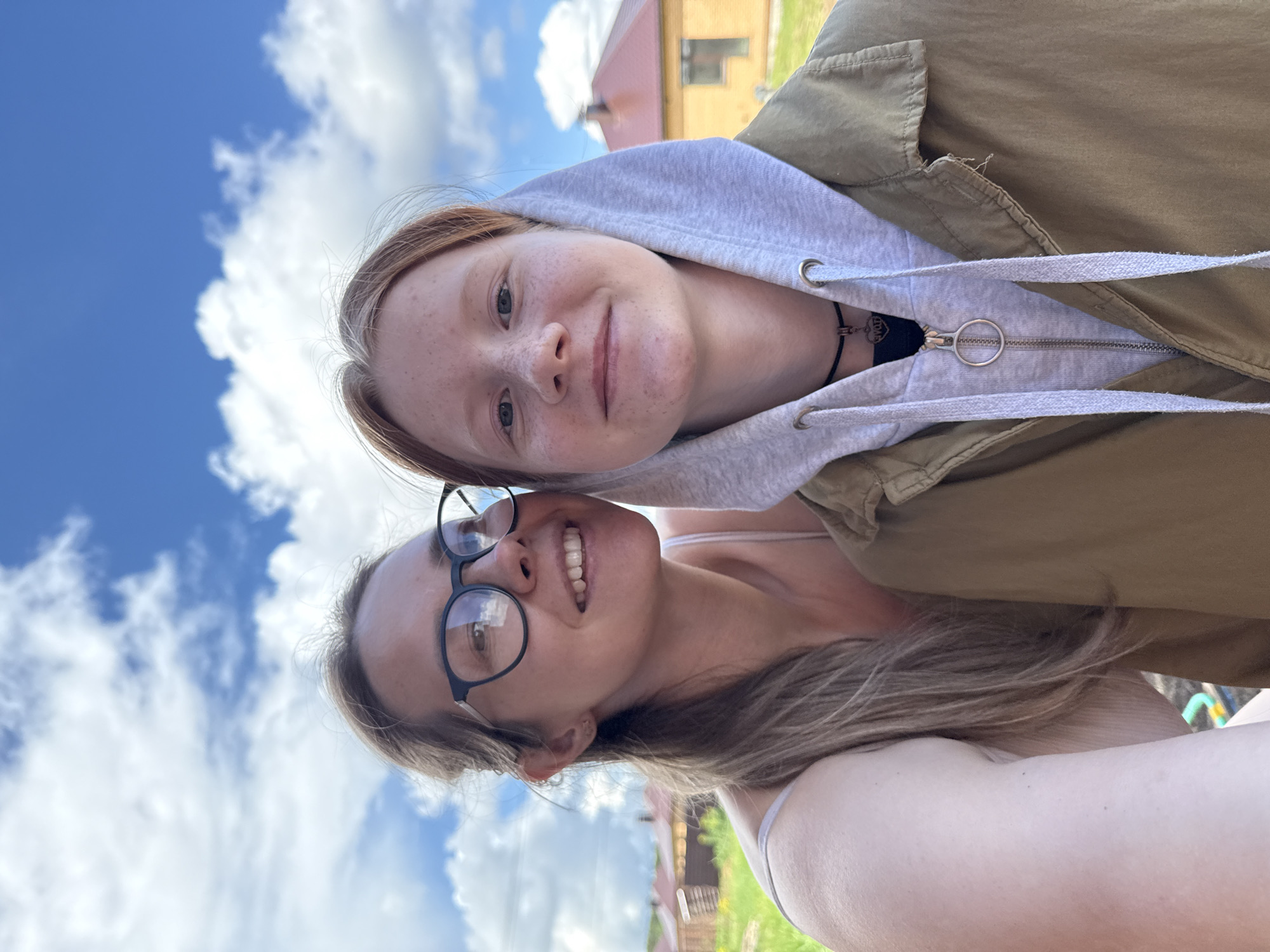 Two women are in the image, seemingly taking a selfie. One woman, positioned lower in the frame, is wearing glasses and has long blonde hair. She is smiling. The other woman is above her, wearing a gray hoodie under a brown jacket. She has red hair and freckles. They appear to be outside. In the background, there is a blue sky with white clouds, a building with a red roof and yellow walls, and some greenery. The location is Chaikovsky, Russia.