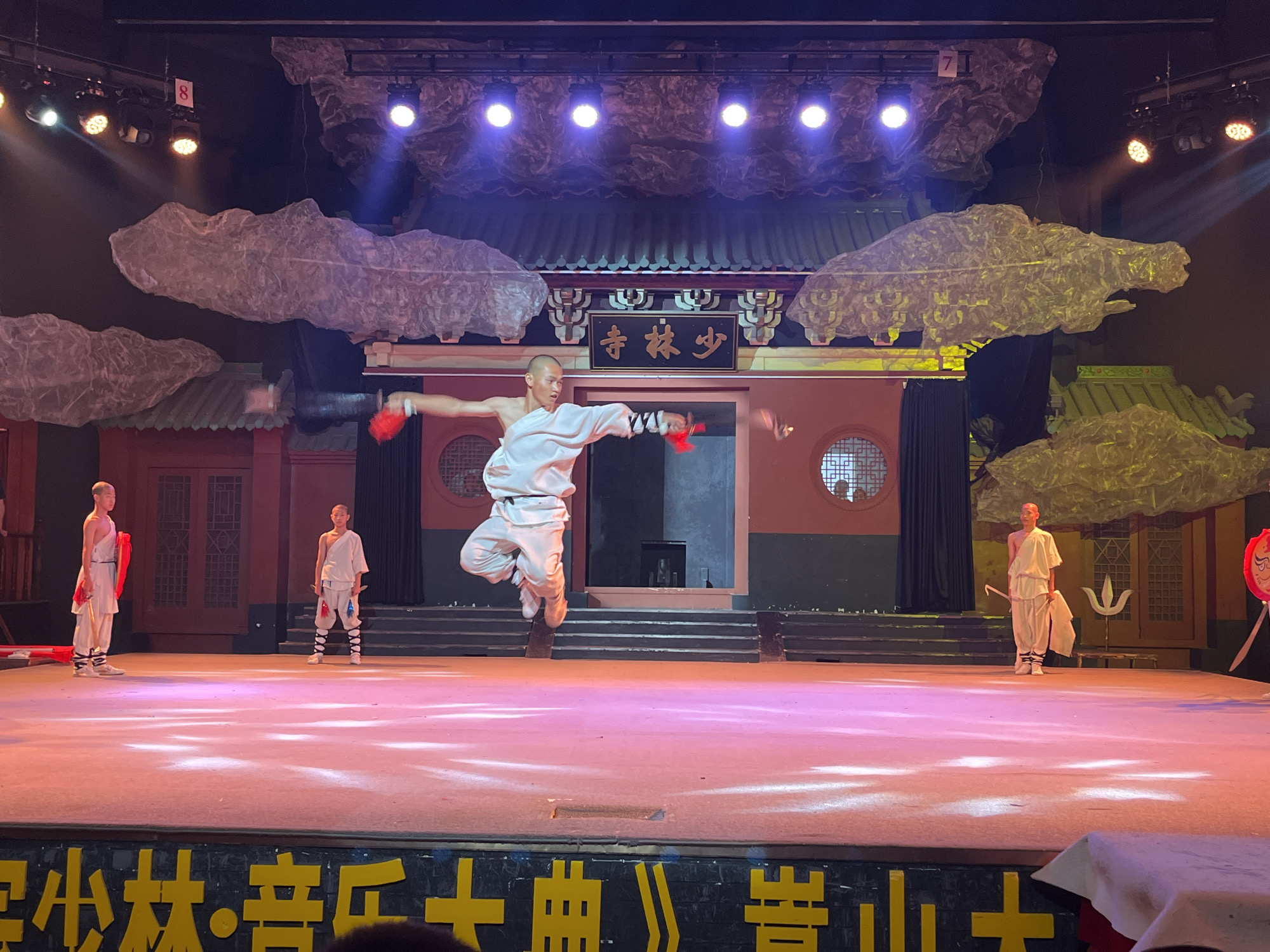 The image depicts a performance on a stage, likely in a theatrical setting. In the foreground, a performer is captured mid-air, in a dynamic jump or kick, wearing a white martial arts uniform. The performer has red objects in each hand, possibly weapons or props. To the left and right of the central performer, other individuals in similar white uniforms are standing on the stage, observing or awaiting their turn. The stage itself is lit with pink and white lights, creating a vibrant atmosphere. Behind the performers, a backdrop simulates an ancient Chinese architectural setting, complete with pagodas and decorative elements. The stage is also adorned with large, cloud-like structures suspended from above. At the very bottom of the image, a section of yellow Chinese characters on a black banner is visible, suggesting the name of the performance or a related inscription. The overall scene suggests a martial arts or cultural performance.