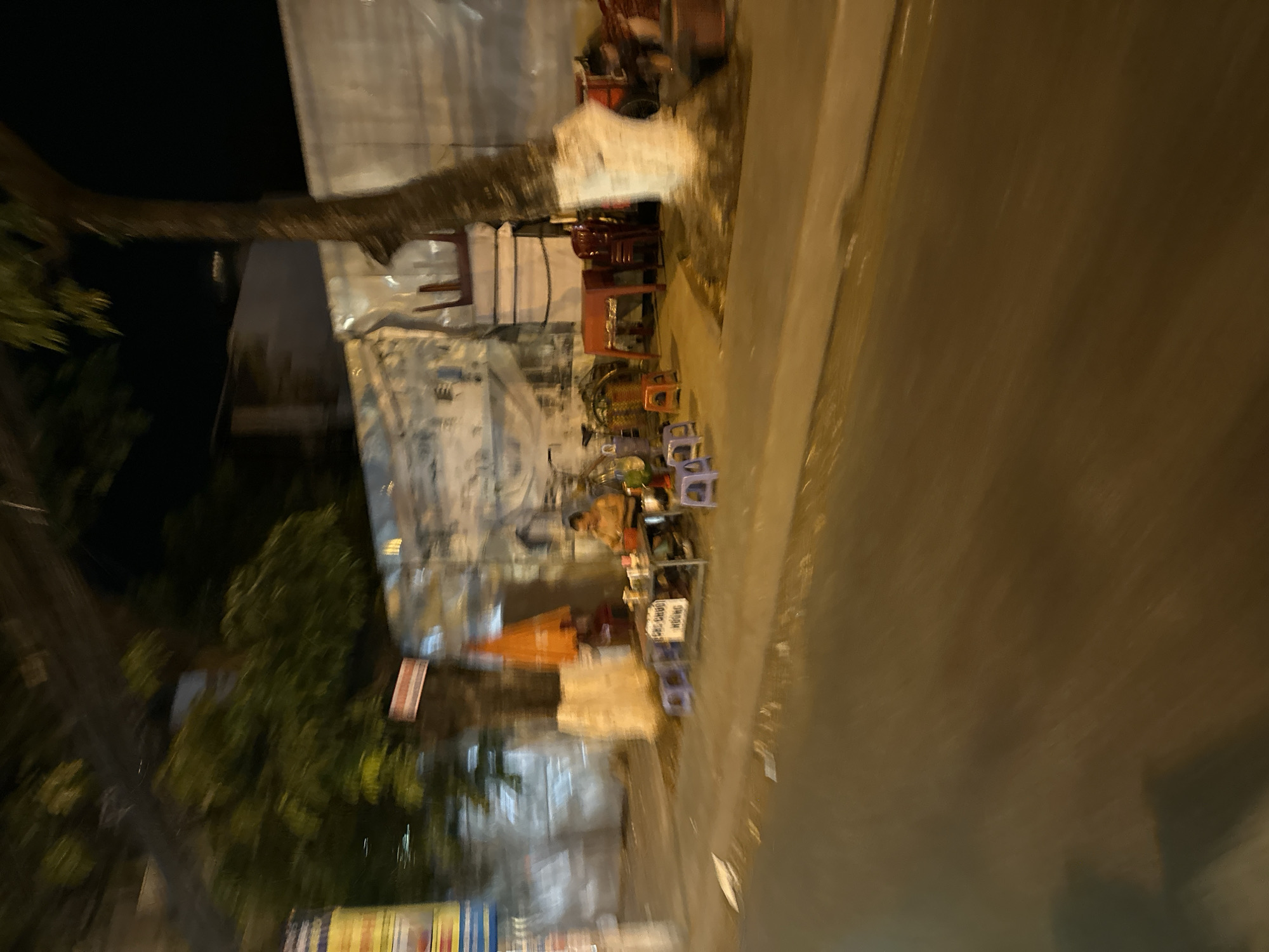 The image depicts a street scene in Bac Nha Trang Ward, Vietnam at night. The view is somewhat tilted. On one side, there's a sidewalk and what appears to be a building or a shop. Tables and chairs are set up on the sidewalk, indicating a street food vendor or a small business operating in the evening. There are also people present, possibly customers. On the other side of the image, there is a street with vehicle traffic. The lighting is artificial, coming from streetlights and the businesses along the sidewalk. The overall atmosphere seems to be typical of a Southeast Asian city street at night.