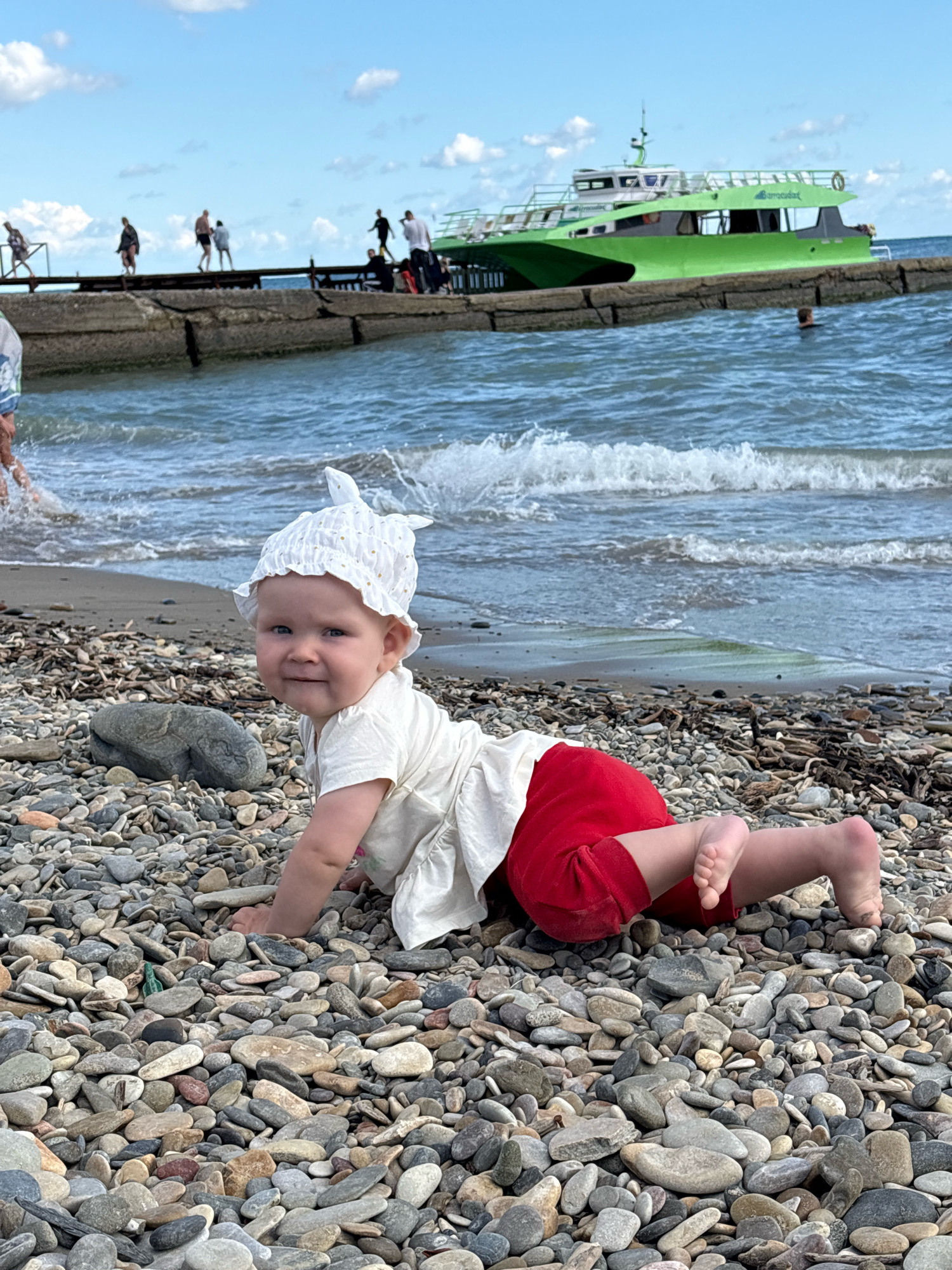 In Arkhipo-Osipovka, Russia, on a pebble beach, a baby girl in a white outfit and a white bonnet is crawling towards the viewer. She is looking at the camera with a slight smile. The sea is behind her, with gentle waves breaking on the shore. In the distance, a pier extends into the water, with several people walking on it. A bright green boat is docked at the pier. The sky is partly cloudy.