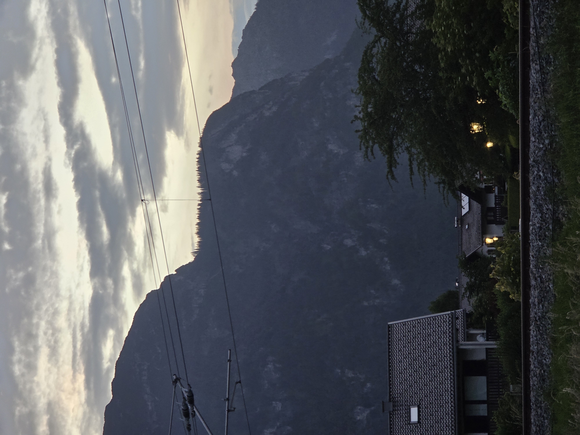 The image depicts a twilight scene in Garmisch-Partenkirchen, Germany, with a mountain range dominating the background. The sky is overcast with textured clouds, illuminated by the fading light. Overhead, electrical wires for a train or tram system are visible. In the mid-ground, several houses with tiled roofs are nestled among trees. Some houses have lights on, casting a warm glow. In the foreground, a railway track runs horizontally across the frame, bordered by gravel and grass. The overall atmosphere is serene and hints at the transition from day to night.