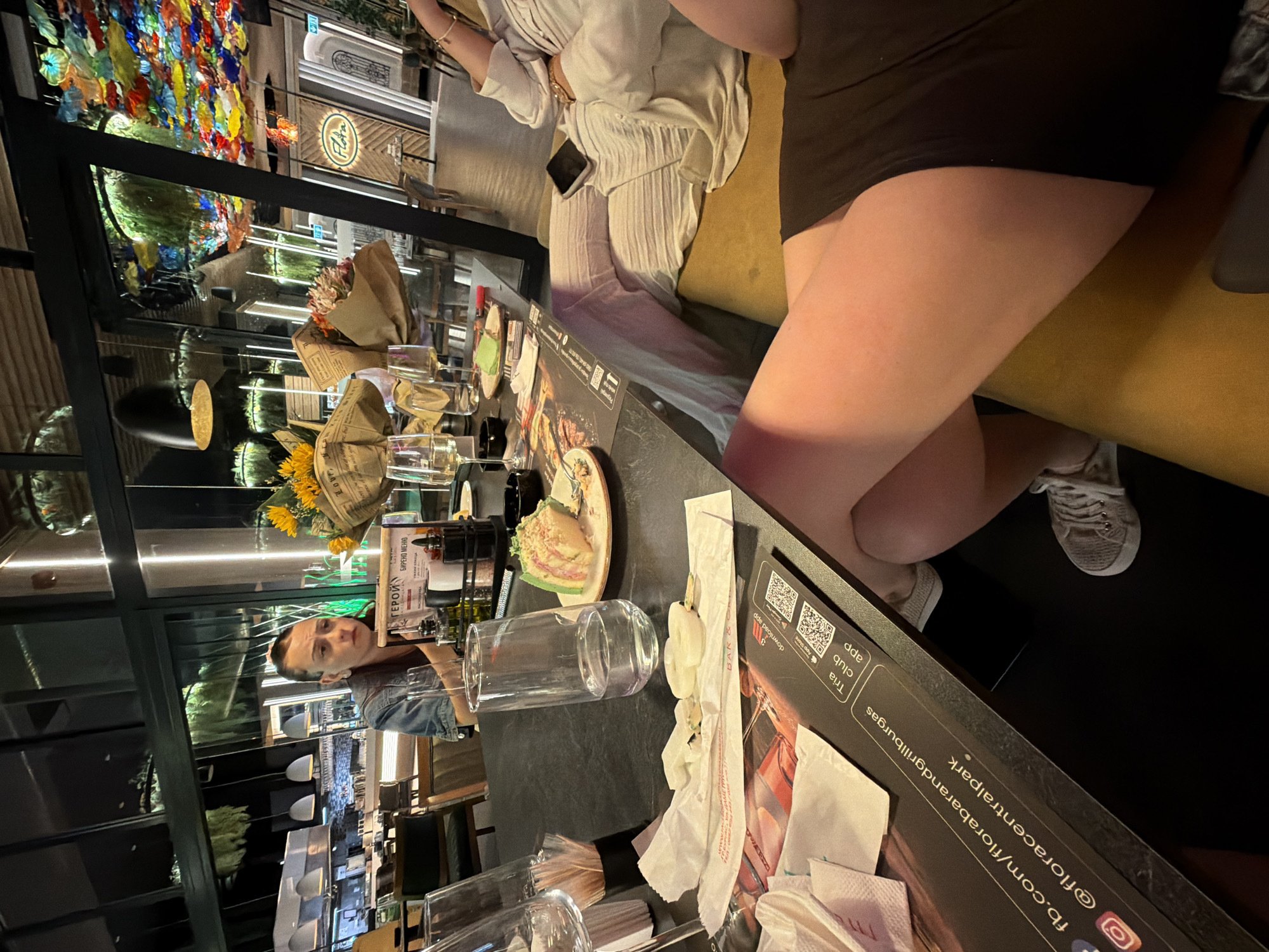 The image depicts a scene at Flora Bar & Grill, located in Burgas, Bulgaria. The setting appears to be a modern restaurant with large windows overlooking an interior space decorated with colorful glass art. The foreground shows a dark tabletop with various items, including plates with remnants of food, glasses, napkins, and a menu with social media handles for Flora Bar & Grill. In the mid-ground, a person is seated at the table, looking down at something. Further back, another person is partially visible, seated on a yellow couch and wearing light-colored pants. The overall atmosphere suggests a dining or social gathering.