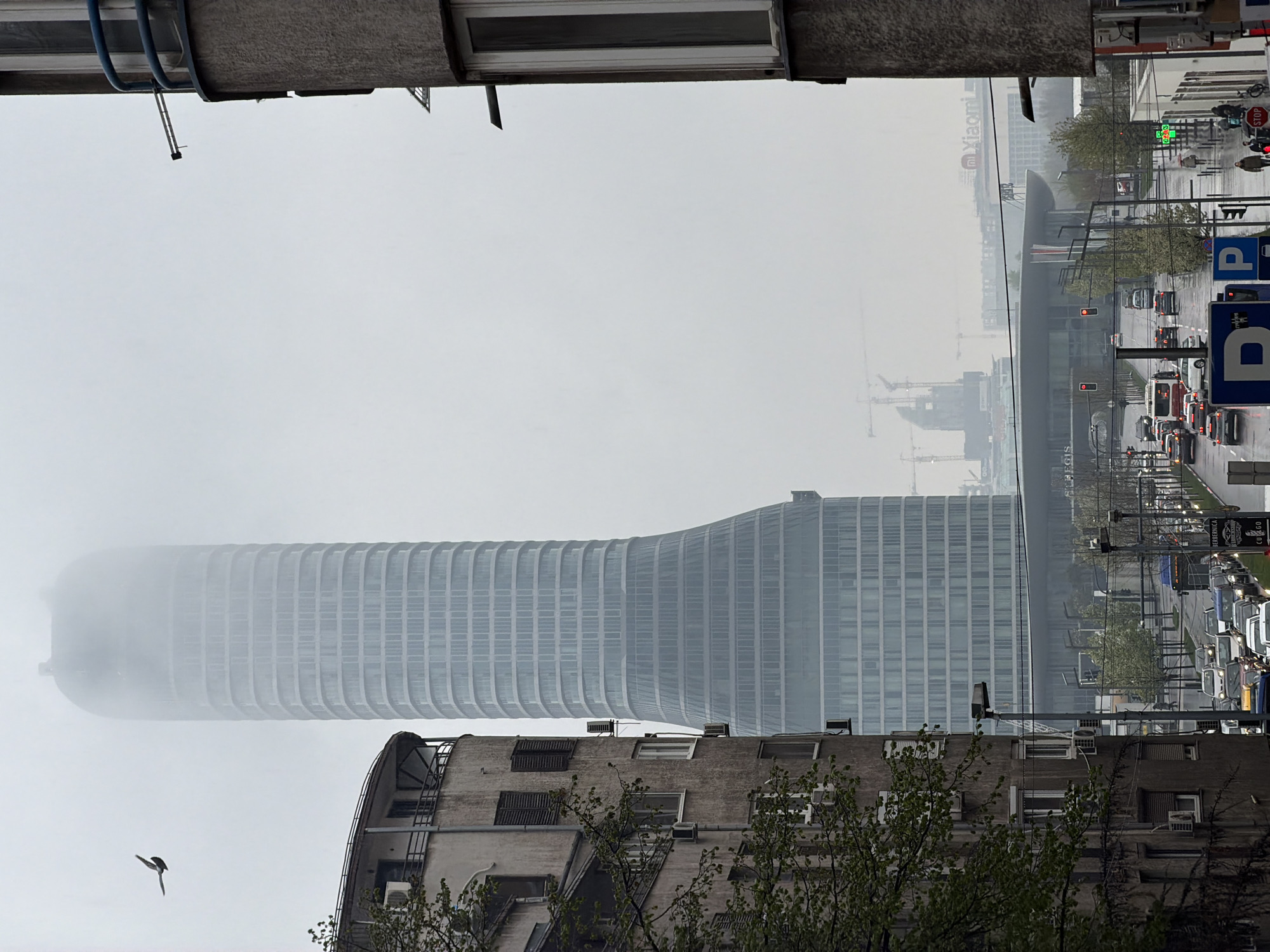 The image shows an upside-down view of a street scene in Belgrade, Serbia, in foggy conditions. A modern, cylindrical skyscraper stands prominently amidst the fog. The skyline is partially obscured by the weather. Buildings are present on the bottom and top of the image. The street scene depicts a traffic-filled avenue with cars, buses, and traffic lights. There are signs with the letter "P" indicating parking. A "Xiaomi" sign is visible on a distant building. A bird is flying in the lower-left part of the image. There are also cranes visible in the background, indicating ongoing construction. The overall atmosphere is urban and somewhat hazy due to the fog.