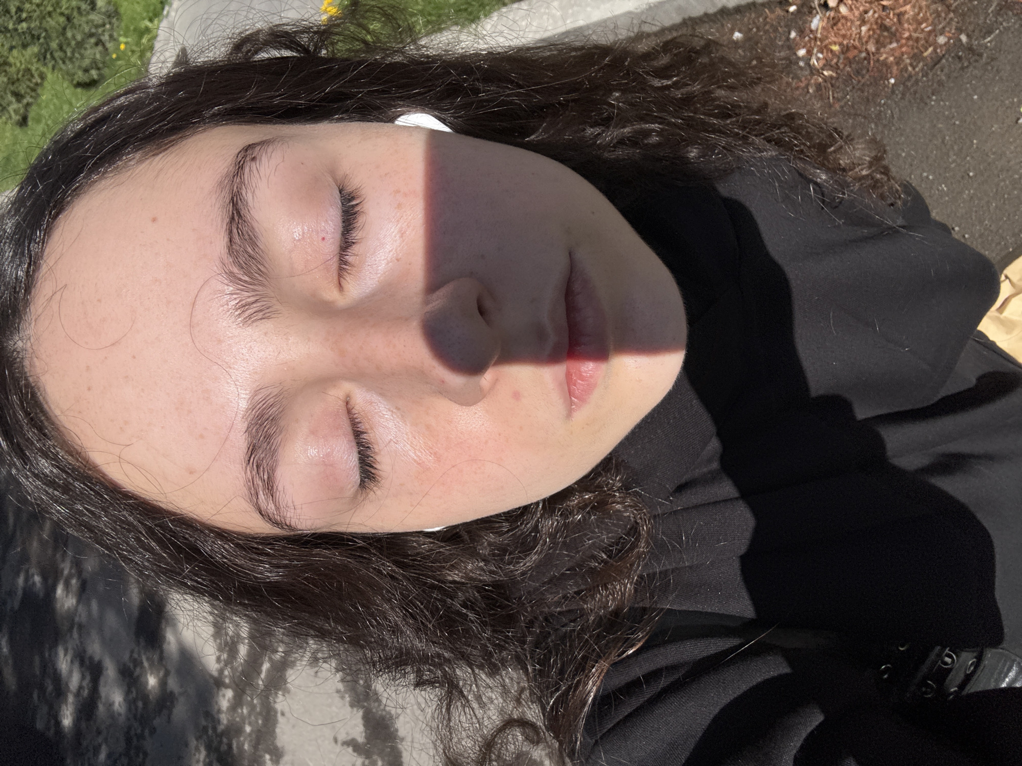 A close-up, slightly off-center photograph shows a person lying down with closed eyes, possibly in a park or outdoor area. The person has fair skin, dark curly hair, and is wearing earbuds. A sharp shadow line cuts across the face. The person is wearing a black hooded top, and the surrounding environment includes greenery and a paved surface with debris. Given the angle of the shot and the presence of grass and foliage, the person may be resting in Yerevan, Armenia, although no specific landmarks confirm this.