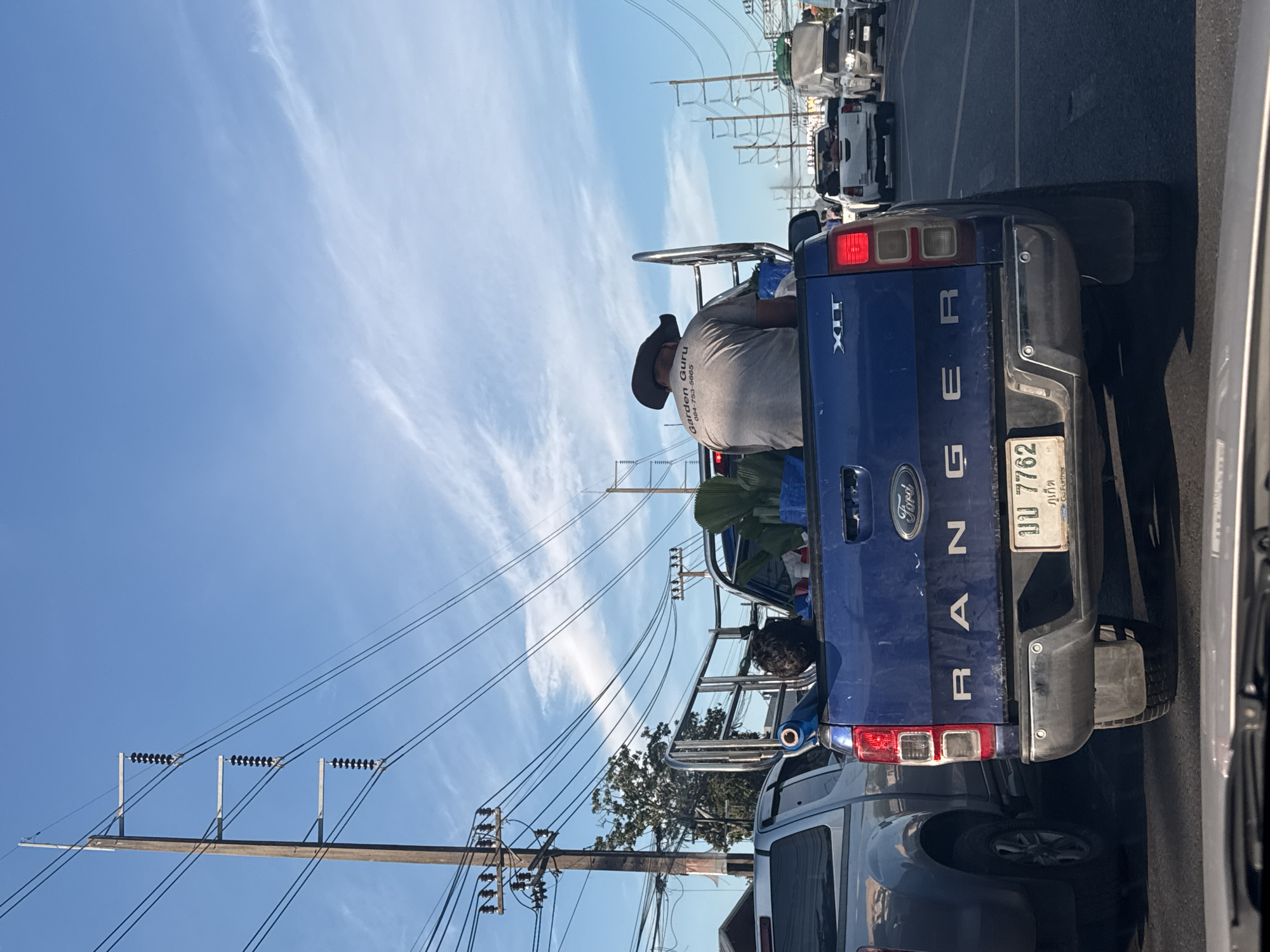A man wearing a cowboy hat sits in the back of a blue Ford Ranger pickup truck, facing away from the camera. The truck is in traffic, with other vehicles visible in front and to the side. The man's shirt has "Garden Guru" and a phone number printed on it. In the bed of the truck, there are some large potted plants. The scene is set against a bright blue sky with scattered clouds and power lines overhead. The license plate on the truck appears to be Thai.