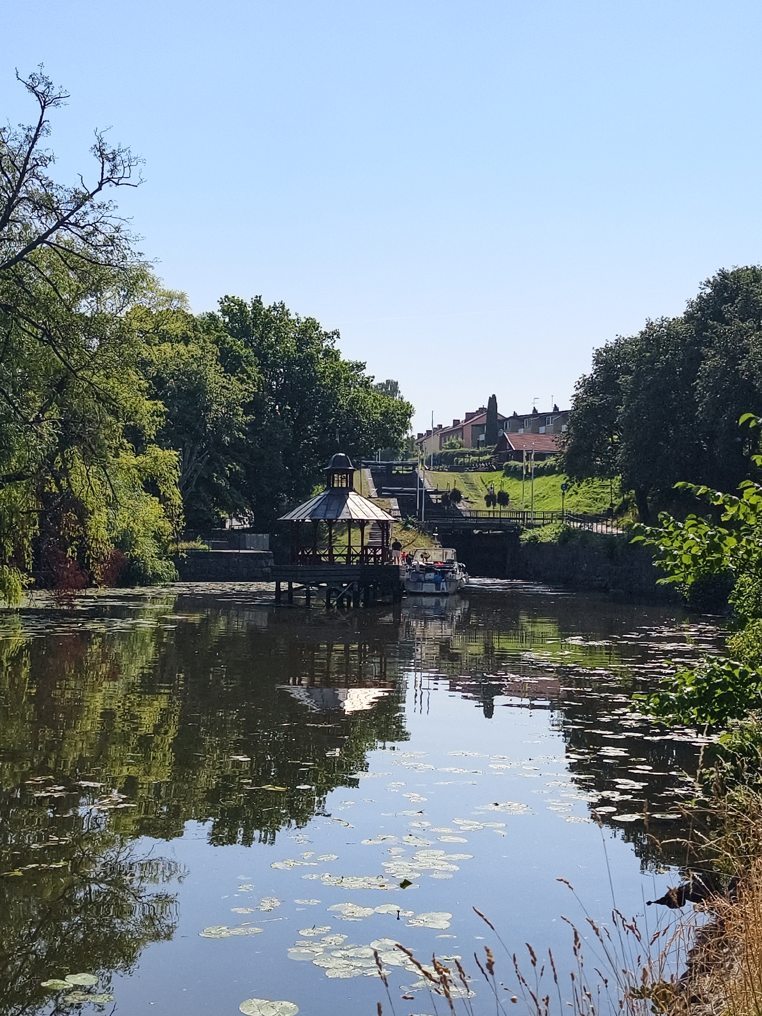 The image depicts a canal or river in Linköping, Sweden, on a clear, sunny day. The water is calm and reflects the surrounding trees and sky. Lily pads are scattered across the surface of the water. On the left side of the frame, lush green trees with some brown foliage overhang the water. In the middle ground, a wooden gazebo with a red roof stands on a pier extending into the water. A boat with several people on board is approaching the gazebo.
Further back, a lock system is visible, with a bridge crossing over it. Beyond the lock, a grassy embankment leads up to a row of residential buildings with red-tiled roofs. The sky is a clear, pale blue. The overall scene conveys a peaceful, natural setting within a populated area.