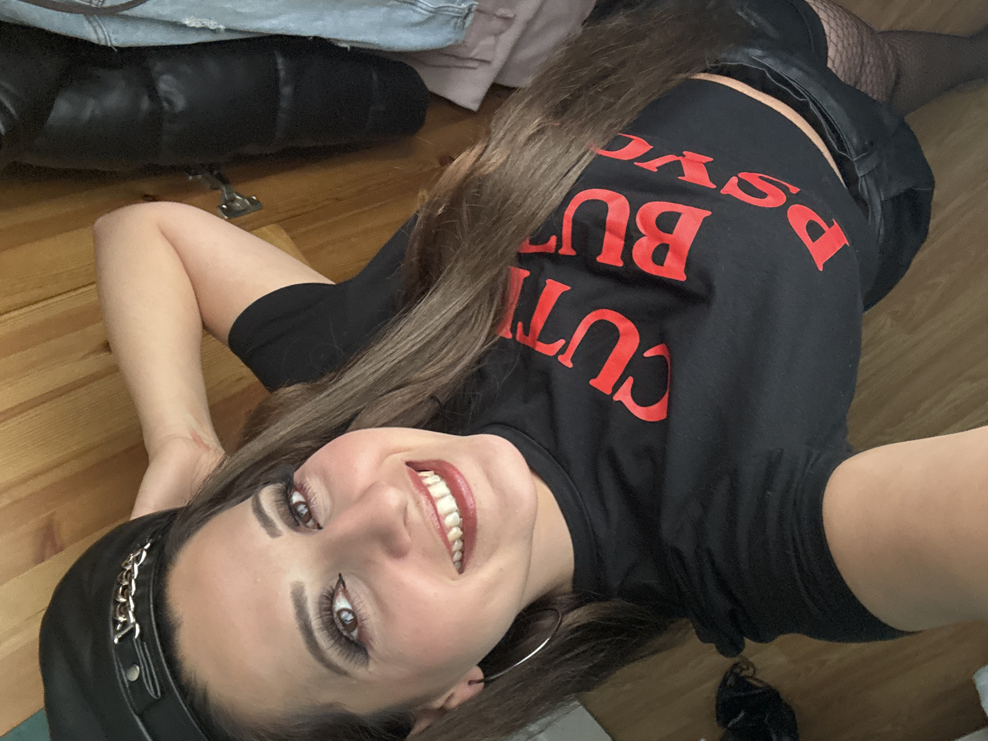 The image shows a woman lying on her back, with her head resting on her arm. She is wearing a black t-shirt with red lettering that appears to say "CURVY BUT PSYCHO", a black leather cap with a chain detail, black shorts, and fishnet stockings. She is smiling and looking at the camera. The background appears to be a wooden surface, possibly a wall or floor. There is no discernible location in the city of Armagh, United Kingdom, as the image does not provide any contextual clues.