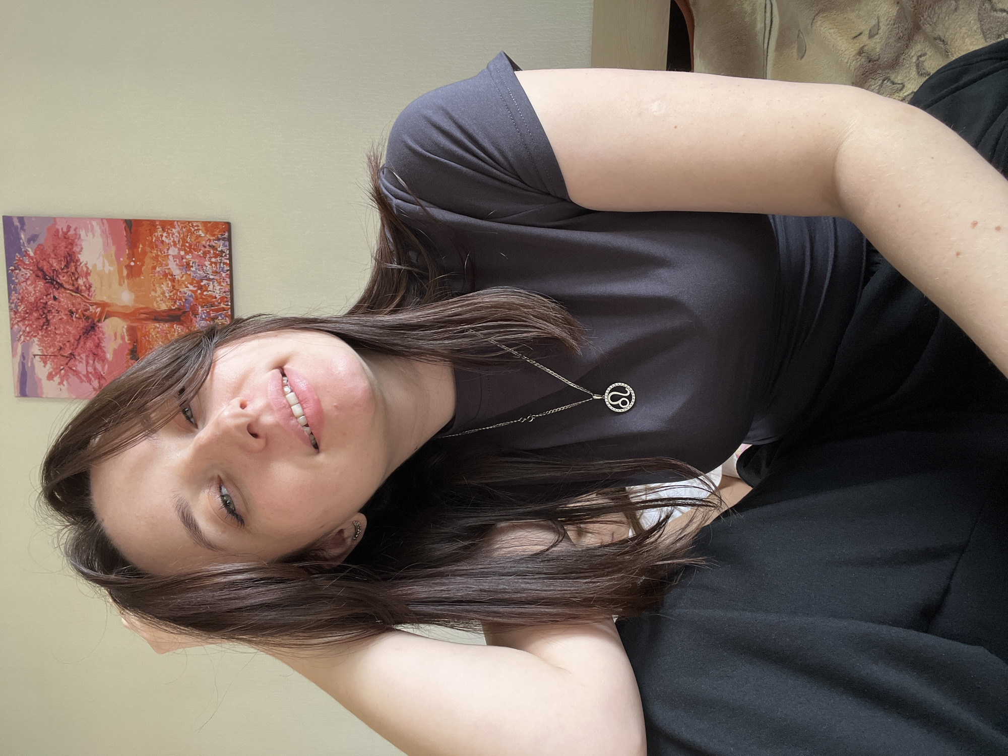 A brunette woman is shown in a medium shot, angled to the left. She is wearing a gray T-shirt and black pants. A silver necklace with a circular pendant is visible. Her hair is long and straight. Behind her, on the wall, hangs a painting of a tree with pink blossoms. A portion of a beige sofa is visible in the upper right corner of the frame. The photograph was taken indoors. There is no information to verify if the photograph was taken in the city of Usol'tseva, Russia.