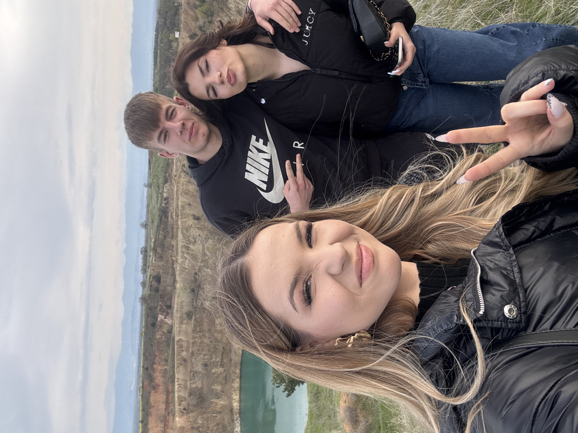A selfie of three young people, taken outdoors in Tsar Asen, Bulgaria. The foreground shows a young woman with blonde hair wearing a black puffer jacket, holding up a hand with two fingers in a "peace" sign. Behind her and slightly to the left is a young man in a black Nike hoodie giving a peace sign as well, standing next to a young woman in a black Juicy puffer jacket with her hand on the young man's shoulder. In the background is a lake bordered by trees, below a cliff face and hills. The sky is overcast.