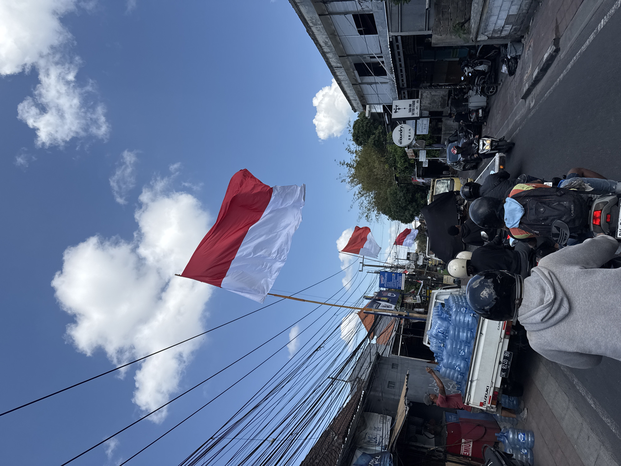 In North Kuta, Indonesia, on a sunny day with scattered clouds, a busy street scene unfolds. The Indonesian flag, red and white, flutters prominently in the foreground, attached to a tall pole. The street is lined with buildings on both sides, many appearing to be shops or businesses, some with metal shutters. A dense network of overhead power lines crisscrosses the sky above the street.
A white Suzuki pickup truck is parked on the side of the road, laden with numerous blue plastic water jugs, suggesting a delivery or supply operation. A man in a red shirt is seen near the truck, possibly involved with the water jugs. Several people are riding motorcycles, the primary mode of transportation, moving along the street. Some riders are wearing helmets, with a few visible from behind, wearing hoodies and backpacks. The road surface is asphalt, and there is a paved sidewalk. The overall impression is of a typical, active day in a bustling urban or semi-urban environment in Indonesia.