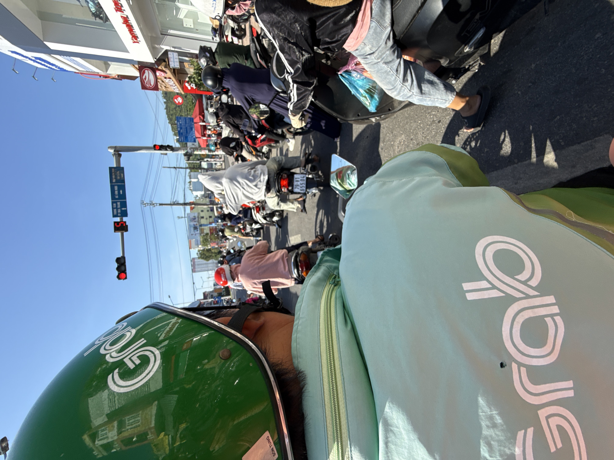 The image captures a busy street scene in Bac Nha Trang Ward, Vietnam, on a sunny day. The perspective is from behind a person wearing a green helmet and a light-colored jacket with a "Grab" logo. The helmet prominently displays the "Grab" logo in white. In the foreground, a portion of the person's light green jacket with a prominent zipper detail is visible, also featuring the "Grab" logo in a pale pinkish-white.
In the middle ground, a traffic light displays a red signal. Numerous motorbikes and their riders are present, indicating traffic congestion. People are wearing helmets and various types of clothing, including hoodies, long-sleeved shirts, and casual wear. Several individuals are wearing masks. One person in the middle ground is wearing a red helmet. A motorbike in the middle ground has a license plate that reads "79-V1 555.04".
The background reveals buildings lining the street, one of which appears to be a commercial establishment with signage. The sky is clear and blue. The overall environment suggests an urban setting with active traffic and pedestrian activity.