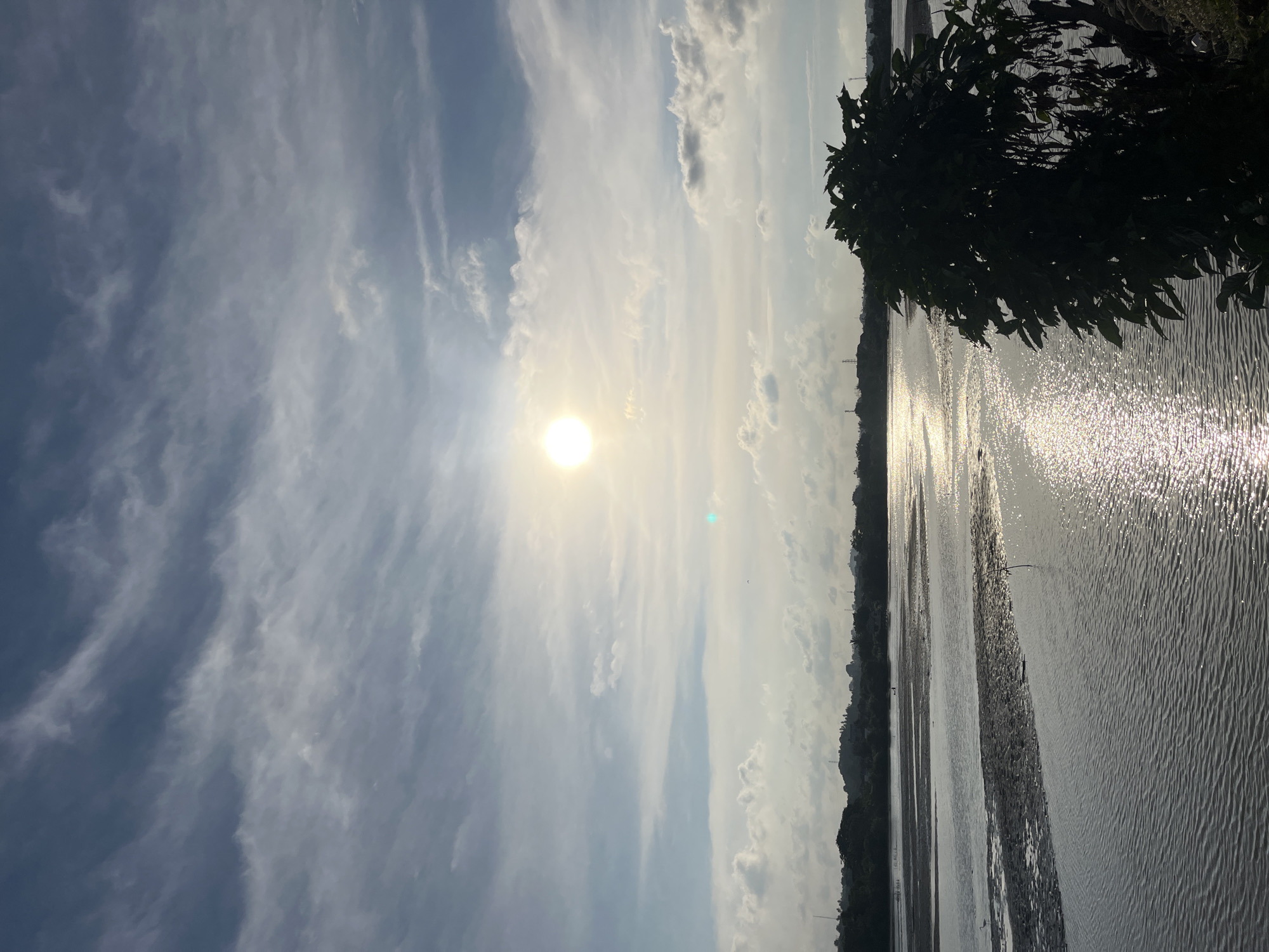 The image shows a body of water with sun glare reflecting off its surface. A dark silhouette of a tree is on the right side of the image. Across the water is a skyline with buildings. The sky is a mix of light blue and white with clouds and the sun shining brightly. The location is likely Lhokseumawe, Indonesia.