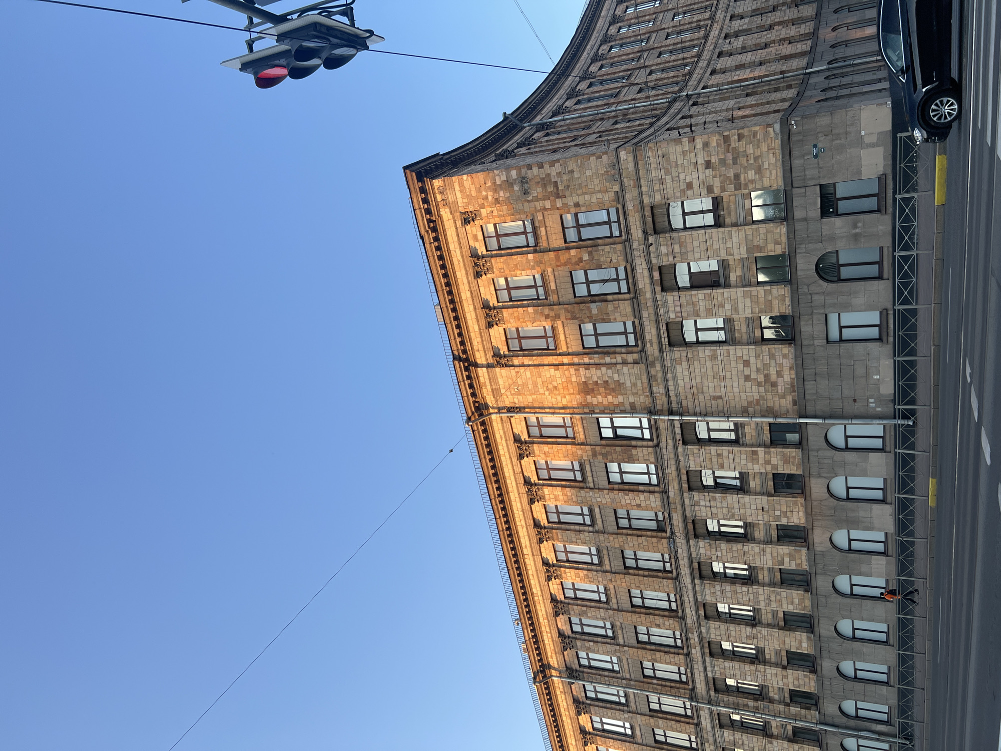 The image depicts an urban street scene in St. Petersburg, Russia, specifically showing the House of City Institutions (Дом городских учреждений) at Mokhovaya Street, 9-11. The large, multi-story building dominates the scene, featuring a light brown, rough-hewn stone facade with classical architectural elements, including pilasters and an ornate cornice. The upper section of the building, particularly the right side, is bathed in warm, golden sunlight, indicating late afternoon or early morning. The lower section of the building and parts to the left are in shadow. A distinctive curved corner defines the building's intersection with another street. Rectangular windows with dark frames are systematically arranged across the facade, while the ground floor features a mix of arched and rectangular windows.
In the foreground, an asphalt road with white dashed lane markings is visible. A low metal barrier separates the road from the sidewalk, where a single person, dressed in an orange top and dark pants, walks from right to left. On the far right, a red traffic light is illuminated, hanging from an overhead pole. Multiple thin, dark wires crisscross the clear, bright blue sky above the building. A dark-colored SUV is partially visible on the right side of the road, heading left. A street sign, partially obscured and illegible due to blur, is visible on the curved corner of the building.