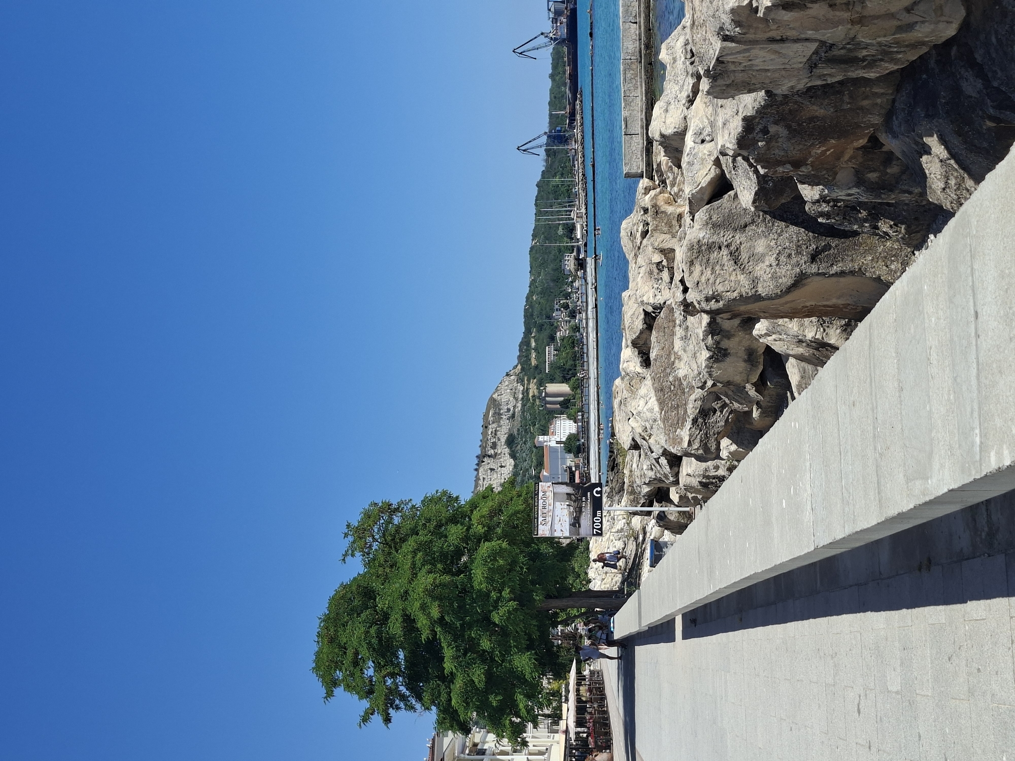 The image depicts a sunny daytime scene at a coastal location. The foreground features a grey concrete walkway and a large, textured rock formation. To the left, a lush green tree stands prominently, with a section of white buildings and outdoor seating visible behind it, suggesting a commercial or residential area.
In the mid-ground, a body of bright blue water stretches out, bordered by a pier or seawall. Several sailboats are docked along the pier, and further in the distance, a hilly landscape is covered in greenery and dotted with buildings. Industrial cranes are visible near the horizon, indicating a port or harbor.
A signpost is placed on the walkway, displaying text that includes "SALT ROOM" and "700m". Two people are seen walking on the walkway near the sign. The overall impression is a pleasant day at a seaside town, with a mix of natural beauty and urban development. The clear blue sky suggests fair weather.