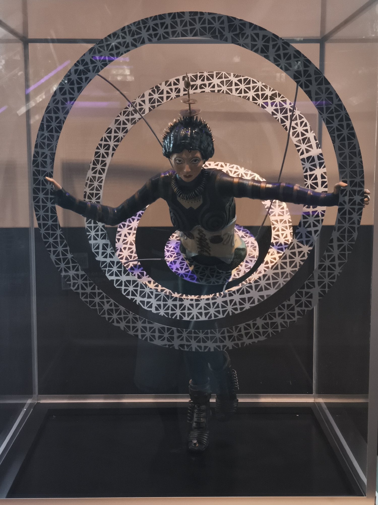 An art display in Barcelona, Spain features a mannequin with an ethnic appearance, inside a transparent display case. The figure wears dark, fitted clothing with light-colored torso details and futuristic-looking footwear. It is surrounded by three concentric, circular frames with patterned designs, which appear to be part of the display. A thin rod extends from the mannequin's head upward, seemingly for decorative purposes. The background behind the display case is dark, with subtle purple lighting. The display case itself is situated on a dark-colored base.
