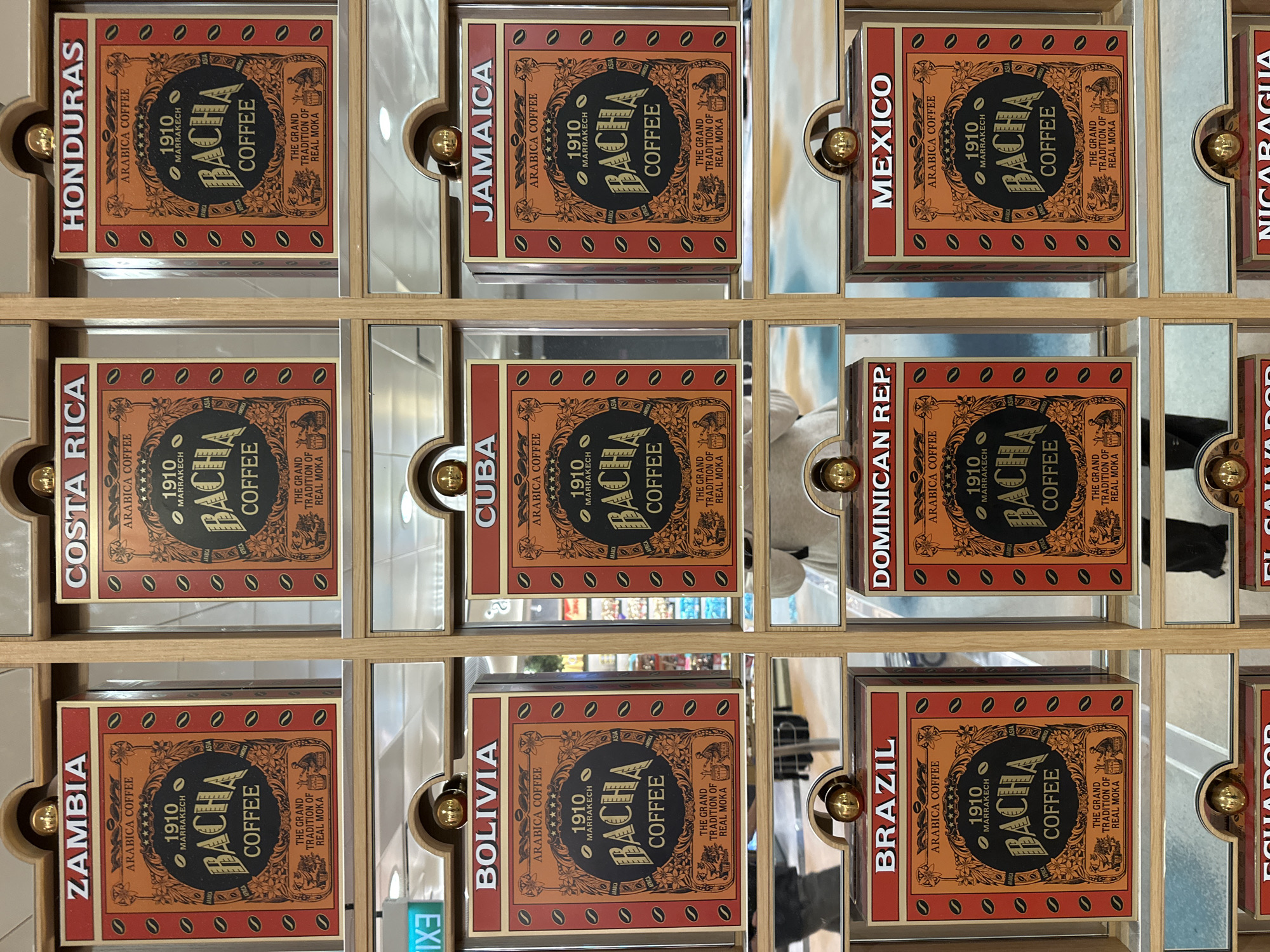 The image displays a retail display featuring numerous canisters of Bacha coffee, each labeled with a country name. The display is divided into a grid of nine visible canisters in the top two rows, with portions of three canisters visible in the bottom row. The canisters are uniformly designed with an orange and black color scheme, featuring the Bacha Coffee logo, "1910 Marrakech," "Arabica Coffee," and decorative motifs.
The visible country names on the canisters are:
Top row, left to right: ZAMBIA, COSTA RICA, HONDURAS.
Middle row, left to right: BOLIVIA, CUBA, JAMAICA.
Bottom row, left to right: BRAZIL, DOMINICAN REP., MEXICO.
The display appears to be a vertical structure with mirrored panels between each set of canisters. The lighting suggests an indoor environment, possibly a retail store or duty-free shop. The reflections in the mirrored panels show parts of a ceiling with fluorescent lights and potentially other display areas or patrons.
One hint image shows a small green sign with "3IX3" visible, which is "EX3" backward and likely part of a larger sign not fully captured.
While the specific location within Singapore is not explicitly stated in the image, the Bacha Coffee brand is known to have retail outlets in Singapore, including at Changi Airport, which is a common location for such elaborate displays. The style of the display and the presence of multiple international brands of coffee are consistent with a duty-free retail environment. No individuals are clearly depicted in focus, though reflections suggest the presence of people.