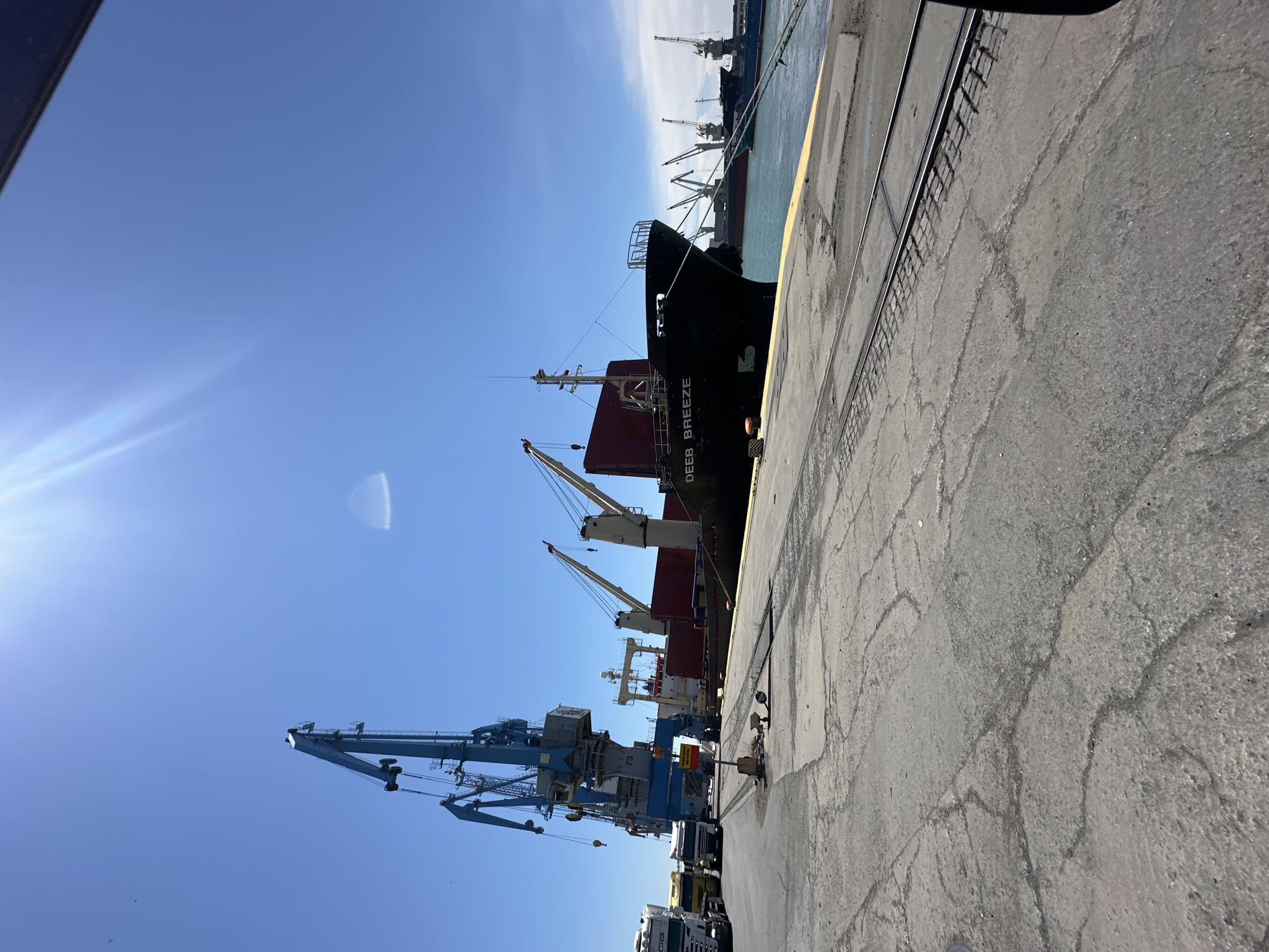 In the city of Unknown, Unknown, on a bright, sunny day, a port scene unfolds. The foreground features a cracked concrete surface with visible train tracks embedded within it, leading towards the water. To the left, a line of white trucks, possibly awaiting cargo, are parked on the asphalt. Dominating the midground is a large cargo ship named "DEEB BREEZE," docked by the water. Two substantial blue harbor cranes are positioned near the ship, with one crane's boom extending over the vessel. In the background, several other cargo ships and cranes are visible, stretching across the tranquil blue water under a clear sky. The sun's glare is prominent in the upper portion of the image.