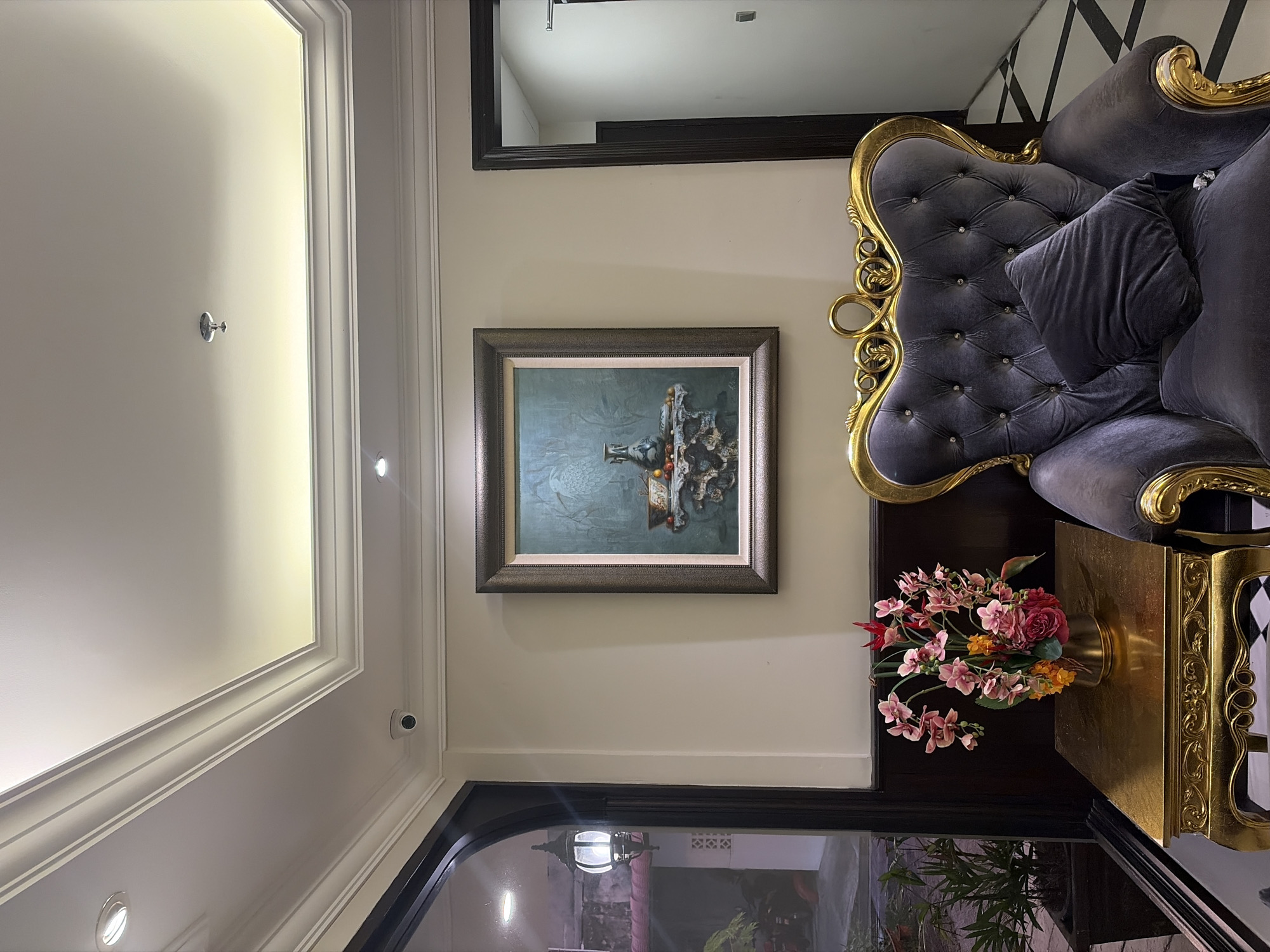 The image depicts an interior scene featuring a luxurious armchair and a golden side table with a floral arrangement. The armchair is upholstered in dark gray velvet with tufted detailing and ornate gold trim. A dark gray cushion rests on the seat. The side table is also golden with intricate carvings. The floral arrangement on the table includes pink and red flowers. Behind the armchair, a framed still life painting is displayed on a neutral-colored wall. To the left of the armchair, a large arched doorway with dark wood trim reveals a glimpse of an outdoor area with a hanging lantern and greenery. The ceiling features recessed lighting and detailed molding. To the right, a doorway leads into a hallway with a patterned floor.