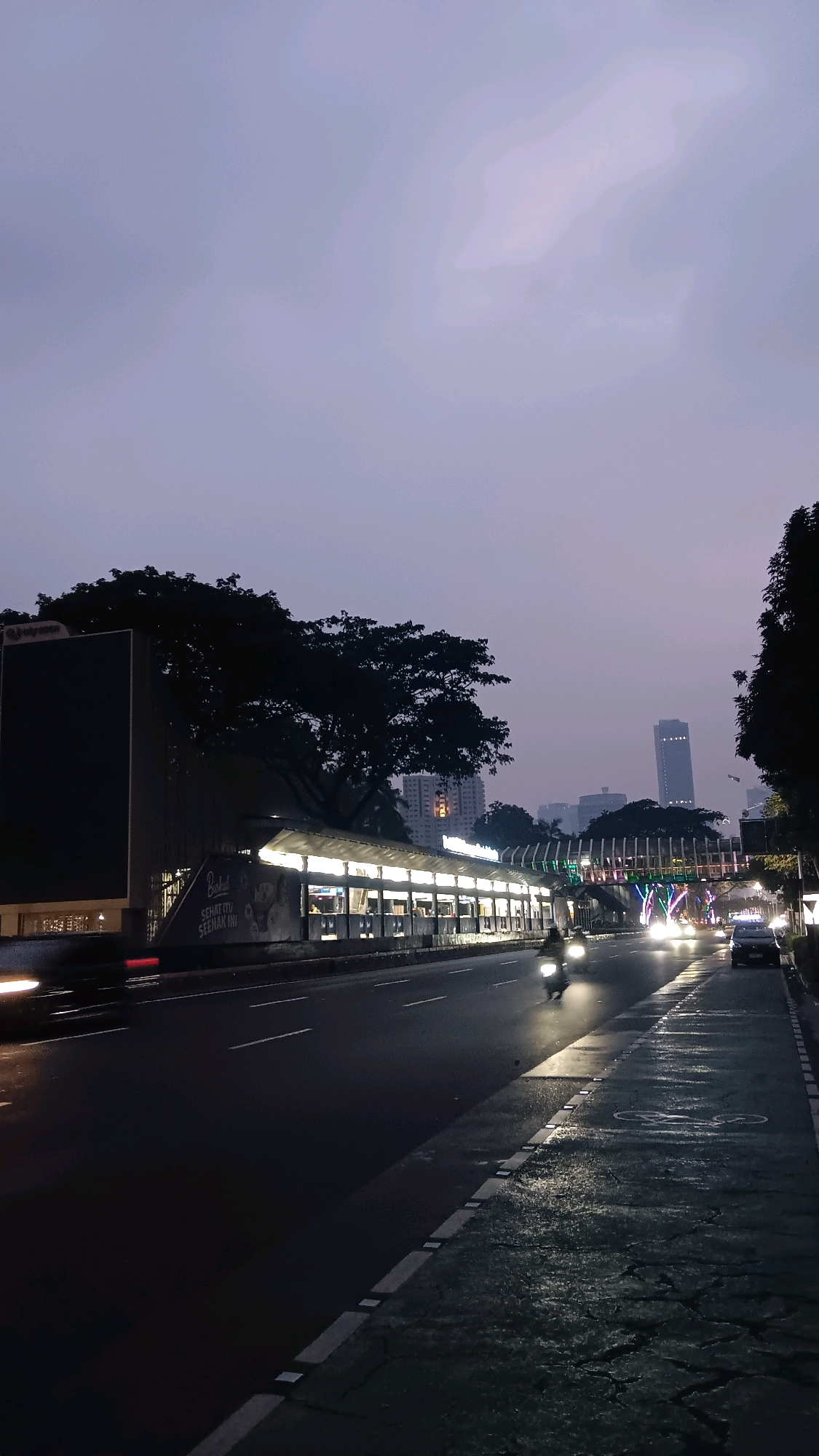 This is a vertical, eye-level, medium shot of a street scene at dusk in South Jakarta, Indonesia. A dark sky with subtle cloud formations hangs above a street lined with trees and buildings. On the left, a large, dark billboard dominates the foreground. Adjacent to it is a covered structure with glowing lights, appearing to be a bus stop or waiting area, with a few indistinct figures inside. The street itself is wet, reflecting the lights of passing vehicles. A motorcycle with its headlight on is moving away from the camera down the street, and other vehicles with illuminated headlights and taillights are visible further down the road. A pedestrian bridge, illuminated with colorful lights, crosses the street in the mid-ground. In the background, several tall buildings rise above the treeline, with the sky transitioning to a lighter shade near the horizon. The overall atmosphere is one of twilight transitioning into night in an urban environment.