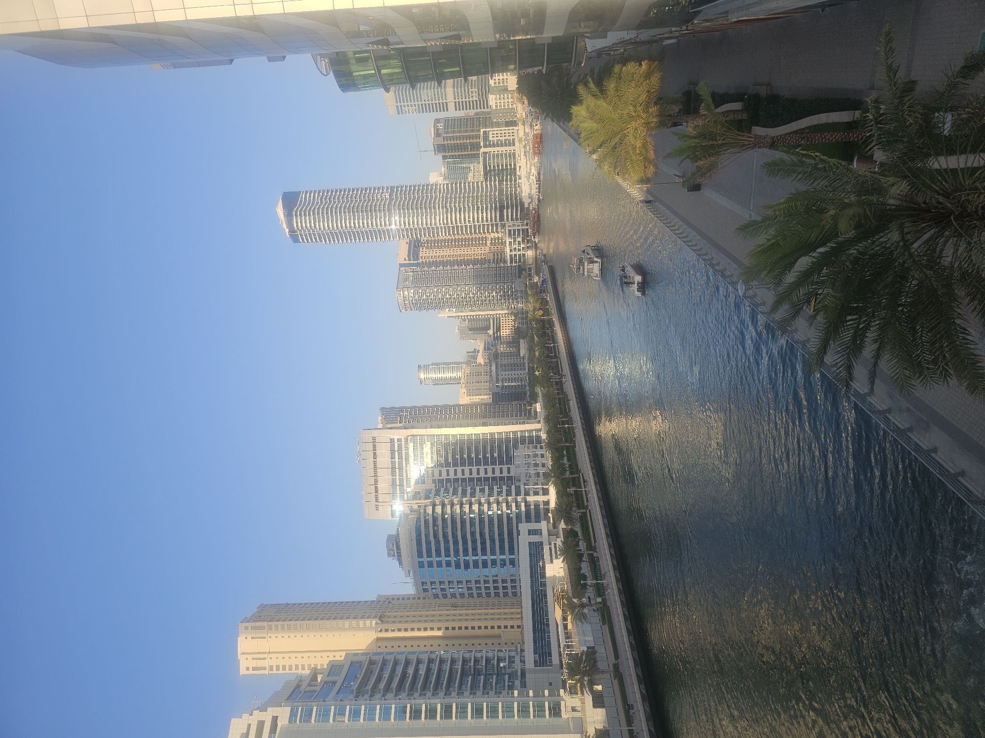 The image shows a cityscape in Dubai, United Arab Emirates. A canal runs through the center of the frame, with boats visible on the water. Tall buildings line both sides of the canal, including skyscrapers and residential structures. Palm trees are interspersed along the canal's edge. The sky is clear and blue. The perspective is from an elevated position.