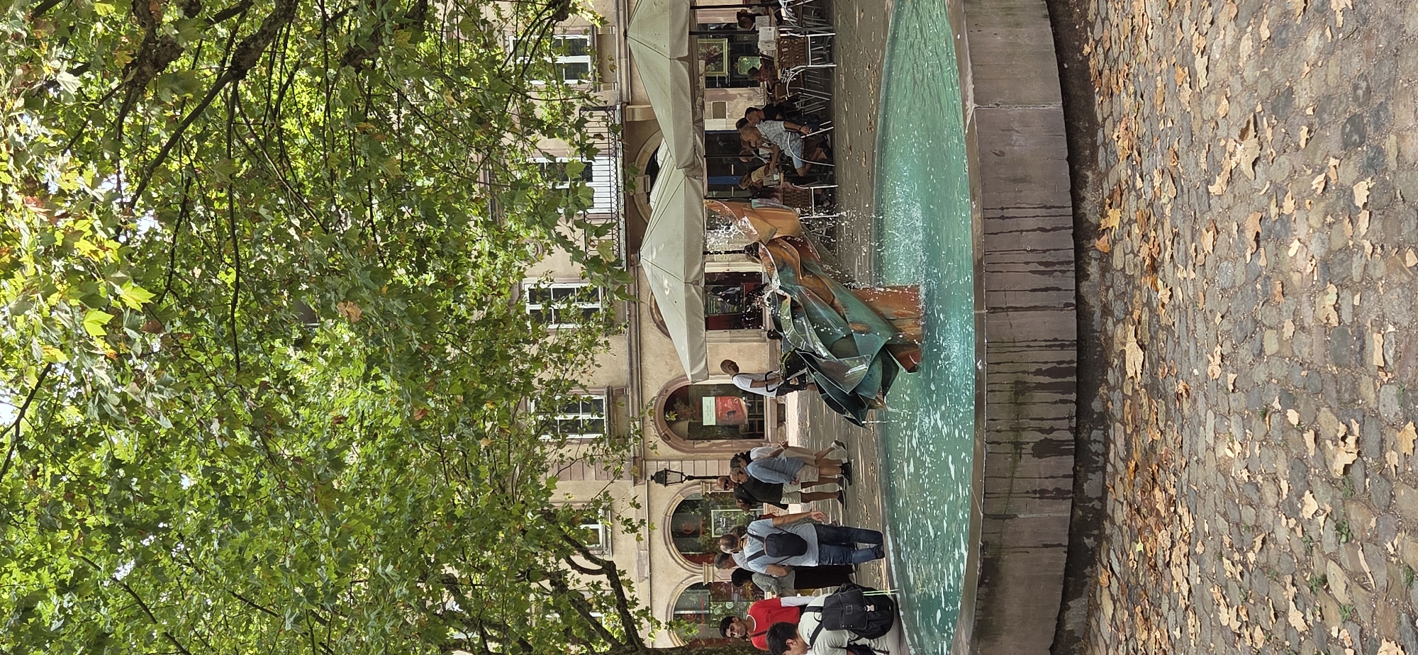 An outdoor fountain with a metallic sculpture is in the foreground. The sculpture is abstract and appears to be made of folded metal in shades of green and brown. Water sprays from the sculpture into the turquoise water of the circular fountain. The fountain's base is made of stone, and the surrounding ground is paved with cobblestones covered with scattered fallen leaves. Behind the fountain, a tree with lush green foliage provides shade, with its branches extending over the scene. In the mid-ground, there are several people walking and sitting at tables under umbrellas, suggesting a cafe or restaurant setting. The background shows a building with arched entrances and windows, characteristic of European architecture. The overall scene depicts a public space in a city, likely a square or plaza.