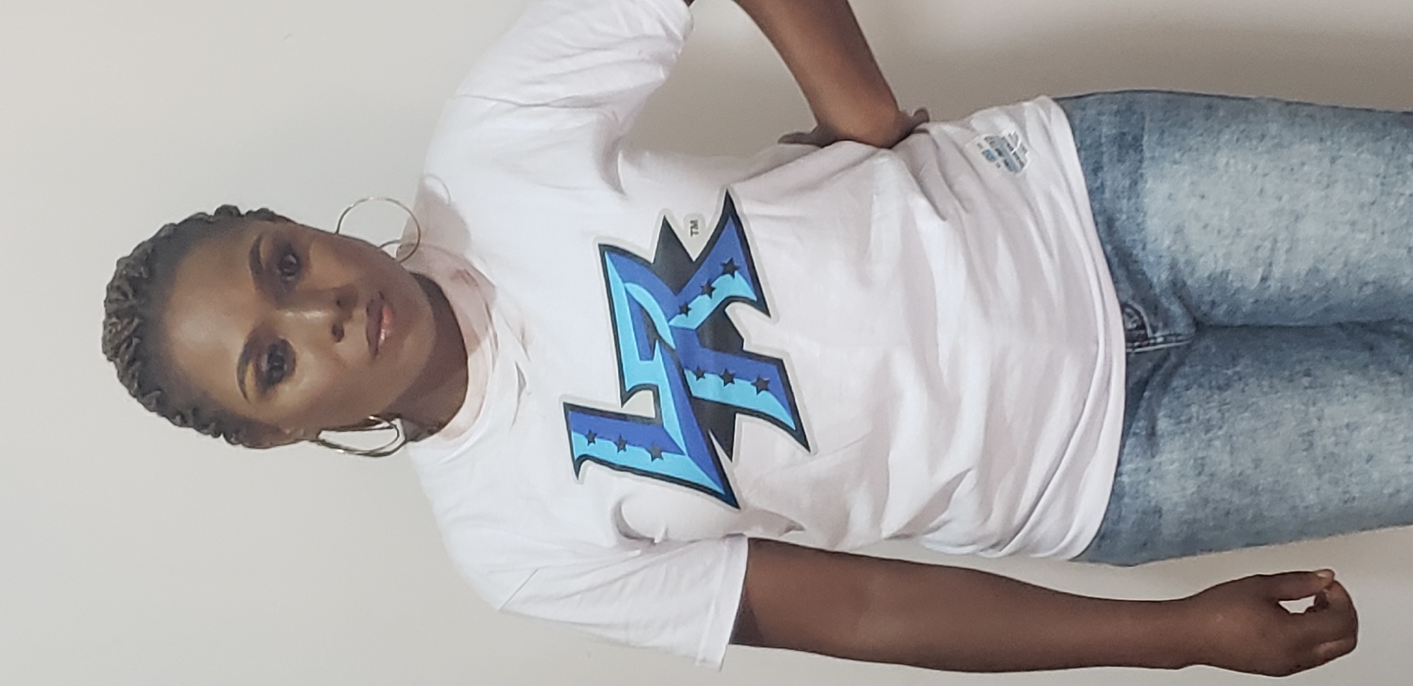 A portrait-oriented, medium shot image shows a woman from the chest up, standing in front of a plain white background. She is wearing a white t-shirt with a blue and black graphic logo, and acid-wash denim jeans. The woman has her right hand on her hip and her left arm is down by her side. She is wearing large hoop earrings and has braided hair. She is wearing eye makeup and lip gloss. The image appears to have been taken in a studio setting. The location is Sabon Gari, Nassarawa, Nigeria.