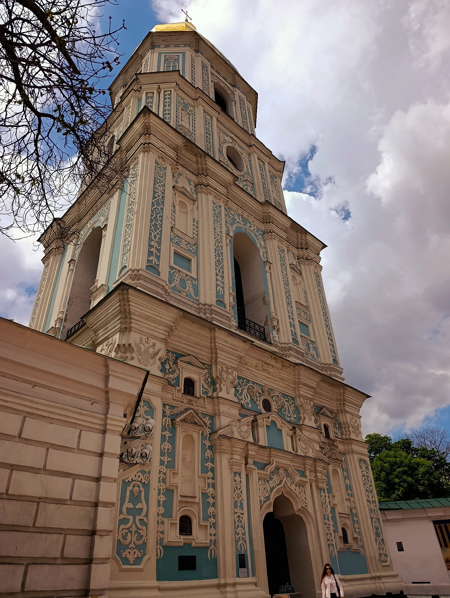 The image captures the bell tower of Saint Sophia's Cathedral in Kyiv, Ukraine, against a partly cloudy sky. The bell tower, a prominent architectural feature, is adorned with intricate white and blue decorative elements. In the foreground, a bare tree branch partially obstructs the view of the tower. To the left of the bell tower is a section of a light-colored wall. At the base of the bell tower, an entrance archway is visible, and a woman wearing a white jacket and sunglasses stands in front of it, facing away from the camera. Green trees can be seen behind the wall to the right of the bell tower. The overall scene depicts a historic religious site under a dynamic sky.