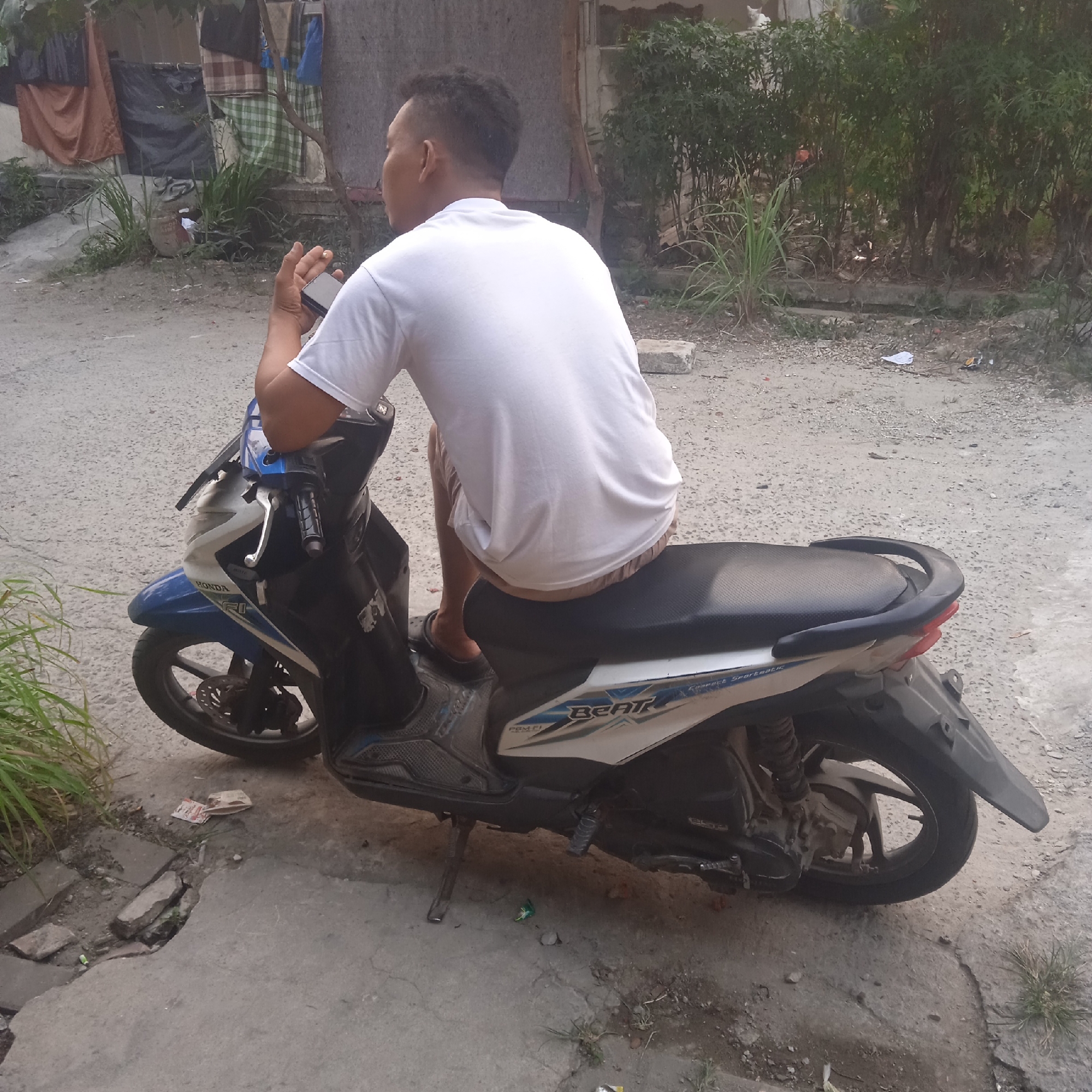 The image depicts a man sitting on a Honda Beat scooter, holding a mobile phone in his right hand. He is wearing a white t-shirt and brown shorts. The scooter is parked on a dirt and concrete ground, likely in a residential area. Behind the scooter, there is a building with laundry hanging outside, and some overgrown vegetation. The overall setting suggests a humble urban environment in Situterate, Indonesia. The ground is uneven, with cracks and debris scattered around. The man's posture and the presence of the scooter indicate he might be taking a break or waiting for someone. The image captures a moment of everyday life in this Indonesian location.