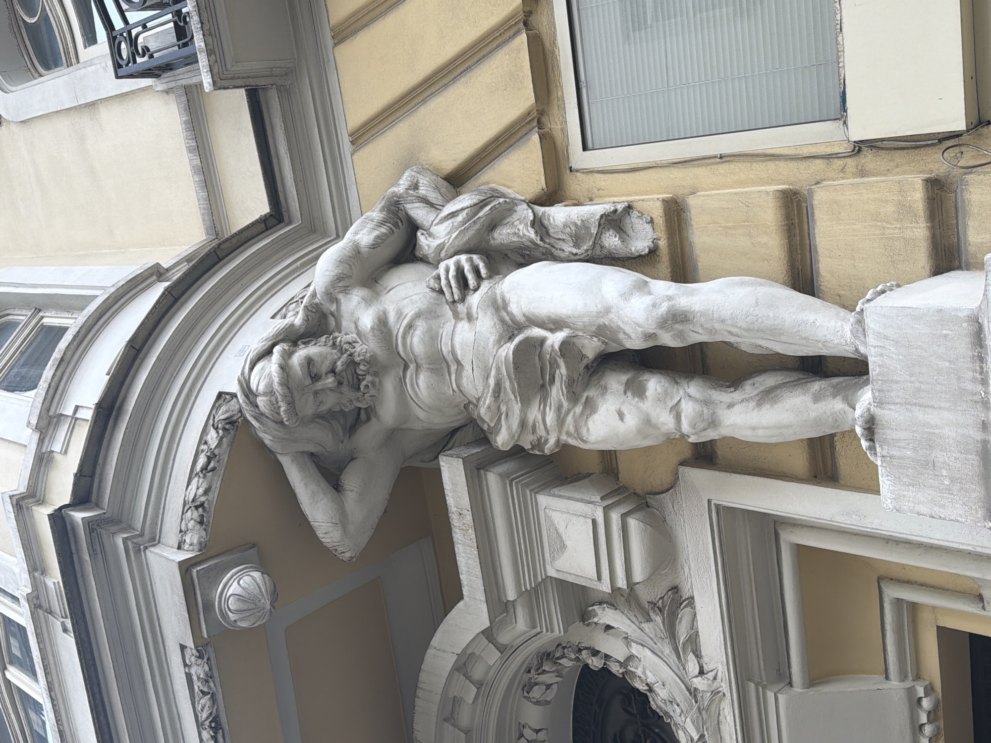The image shows a white statue of a reclining male figure adorning a building facade in Sofia, Bulgaria. The statue is positioned above a doorway or archway and appears to be made of stone or a similar material. The building has a light yellow facade with decorative architectural elements. A window is visible above the statue.