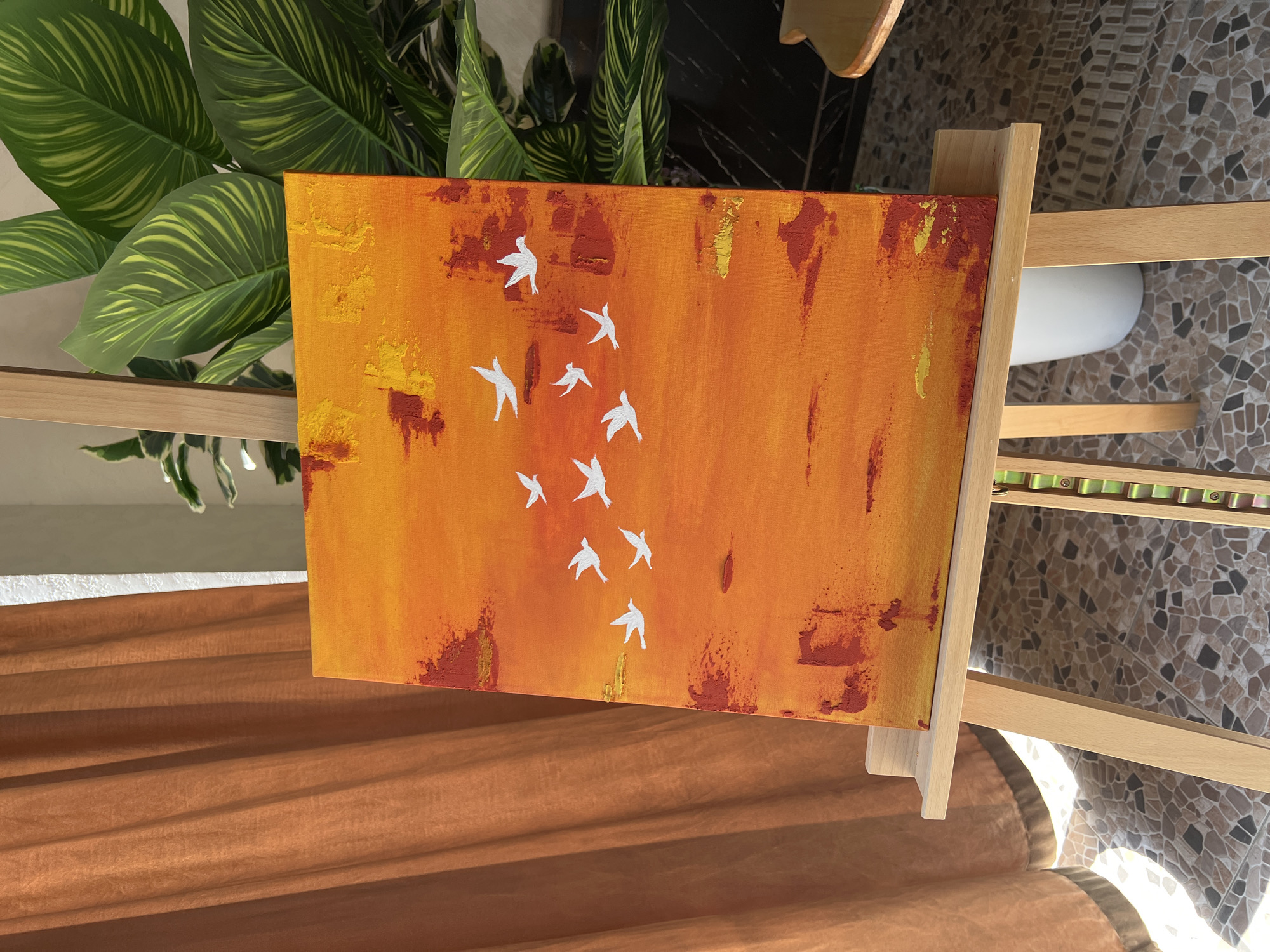 An abstract painting of white birds in flight against a fiery orange and yellow sky is displayed on a wooden easel. The painting features textured brushstrokes of reds and oranges, creating a vibrant, sunset-like backdrop. A flock of approximately ten small, white birds is depicted in various flying poses, scattered across the canvas. The easel is positioned in front of a backdrop of earthy-toned curtains on the left and lush green plants on the right. The floor is tiled with a mosaic of brown and beige stones. The overall scene suggests an indoor setting, possibly an art studio or a well-lit room, with natural light casting shadows. There is no indication of the city of Namangan City, Uzbekistan, within the image.