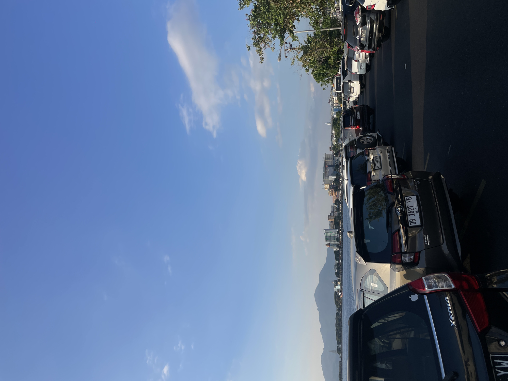 The image shows a parking lot filled with cars next to a body of water, with a city skyline and mountains in the background. The parking lot appears to be located in the city of Manado, Indonesia, as indicated by the license plates on the cars (e.g., "DB 1627 YB," "DB 1169 DJ," "DB 1342 LC," which are Indonesian license plates from the Sulawesi region). The sky is blue with scattered white clouds. The water is calm, and there are buildings and trees visible on the far shore. The cars are parked in rows, facing away from the viewer. The overall scene suggests a coastal urban environment.