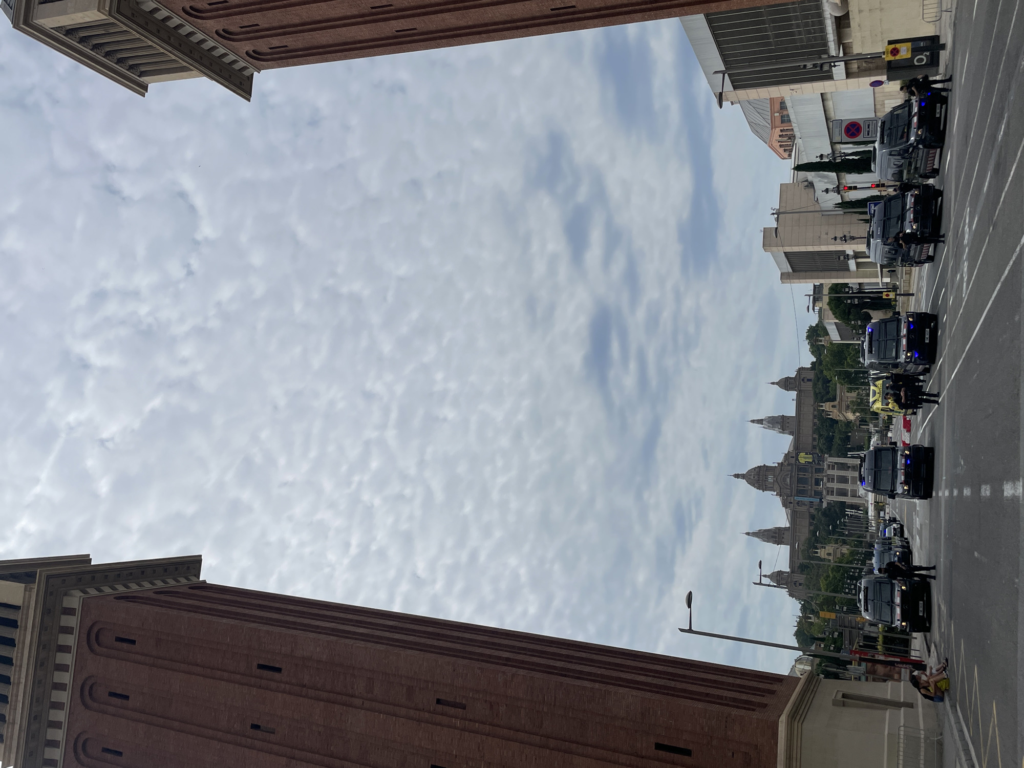 This image captures a street scene in Barcelona, Spain, likely near the Palau Nacional on Montjuïc hill. In the foreground, two imposing brick towers flank the composition, characteristic of the Venetian Towers that mark the entrance to the National Art Museum of Catalonia. A street runs between them, lined with several black vans, some appearing to be police vehicles, with blue flashing lights visible on some. Several individuals, some in uniform, are present around the vans. A few civilians are seated on the sidewalk on the left side of the street. In the background, the grand facade of the Palau Nacional, with its distinctive domes and towers, is visible under a cloudy sky. The overall atmosphere suggests a controlled or heightened security presence in a public space.