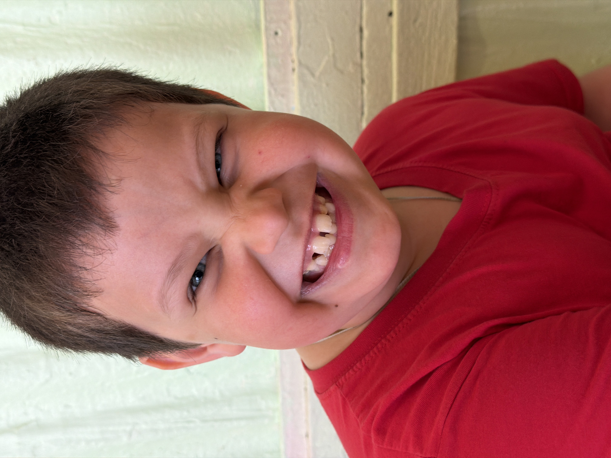 A close-up, Dutch-angled portrait shows a young child, likely a boy, from Izhevsk, Russia, looking directly at the viewer with a broad, joyful smile. He has short, dark brown hair and expressive blue eyes that are crinkled with amusement. His mouth is wide open, revealing his teeth, which include both new permanent teeth and gaps from lost baby teeth, characteristic of a child his age. A slight discoloration or shadow is visible on some teeth.
The child is wearing a plain red t-shirt, and a thin, light-colored chain is visible around his neck. His fair skin shows a few minor blemishes on his cheek and nose area. The background features a light-colored wall, possibly off-white or very light green, with vertical elements that suggest paneling or wooden beams, indicating an indoor or sheltered domestic setting. The lighting is bright and even, highlighting the child's cheerful expression. The image conveys a playful and happy moment. No text is visible in the frame.