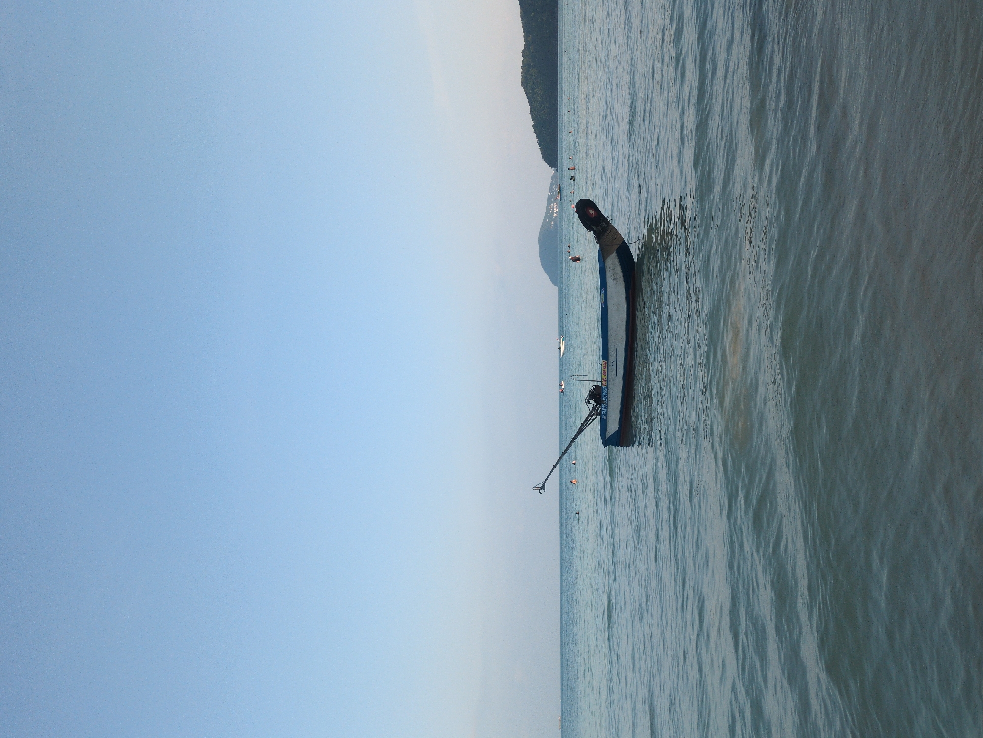 The image shows a partially submerged longtail boat in the shallow waters off a beach in Phú Quốc, Vietnam. The boat is positioned at an angle to the shore, with its bow pointing towards the beach. The water is slightly choppy, and there are other boats and people visible in the distance. A forested coastline is visible on the horizon. The sky is a light blue color.