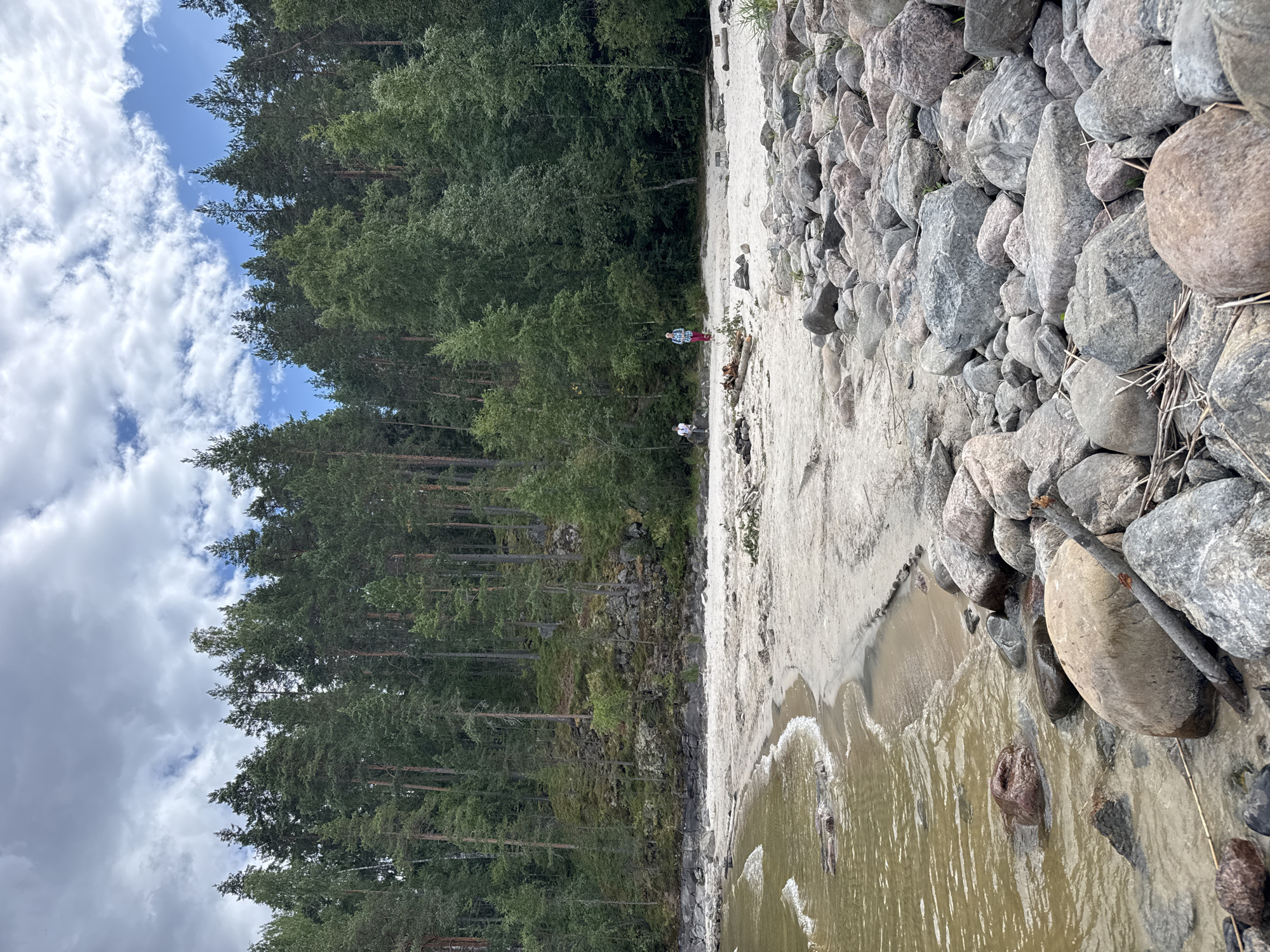 The image depicts a shoreline in the city of Sorola, Russia, characterized by a sandy beach, rocky embankment, and a dense forest. In the foreground, a collection of large, rounded stones covers the right side of the frame, with the brownish water of a lake or river lapping at the sandy shore on the left. The sand stretches back to a treeline, featuring a mix of pine and deciduous trees. Two individuals are visible in the mid-ground on the sandy beach, one standing and looking towards the water, and another walking away from the camera. Scattered campfire pits and some driftwood are also present on the sand. The sky above is partly cloudy, with patches of blue sky visible between white cumulus clouds. The scene appears to be a natural, undeveloped area, likely a popular spot for recreation.