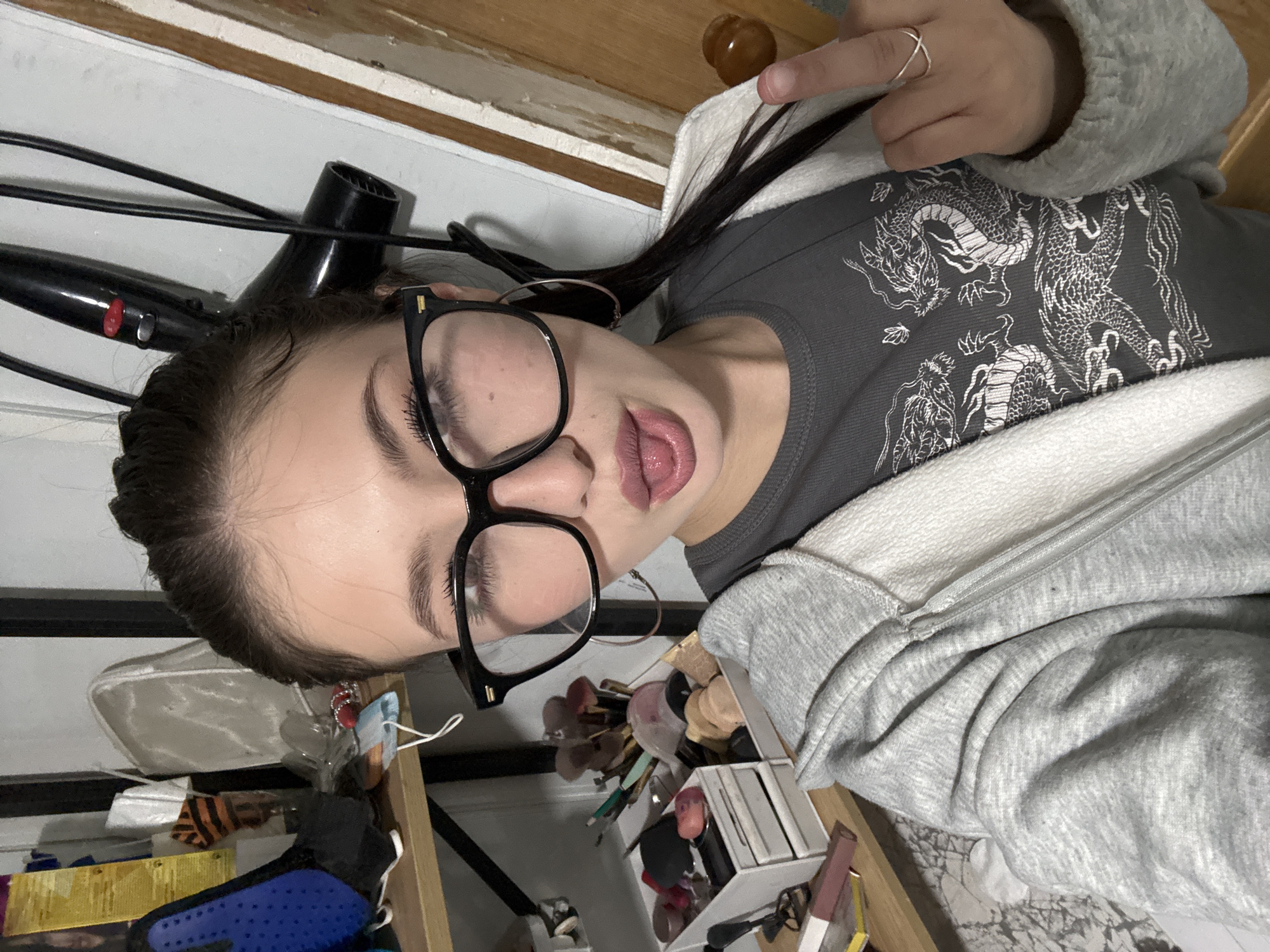 A young woman is lying on her side, possibly on a bed or couch, with a drawer of makeup and a hair dryer in the background. She is wearing glasses, a gray top with a dragon design, and a gray zip-up hoodie. She is sticking her tongue out and giving the middle finger. The location in the city of Фоки, Russia is not discernible from the visual information provided.