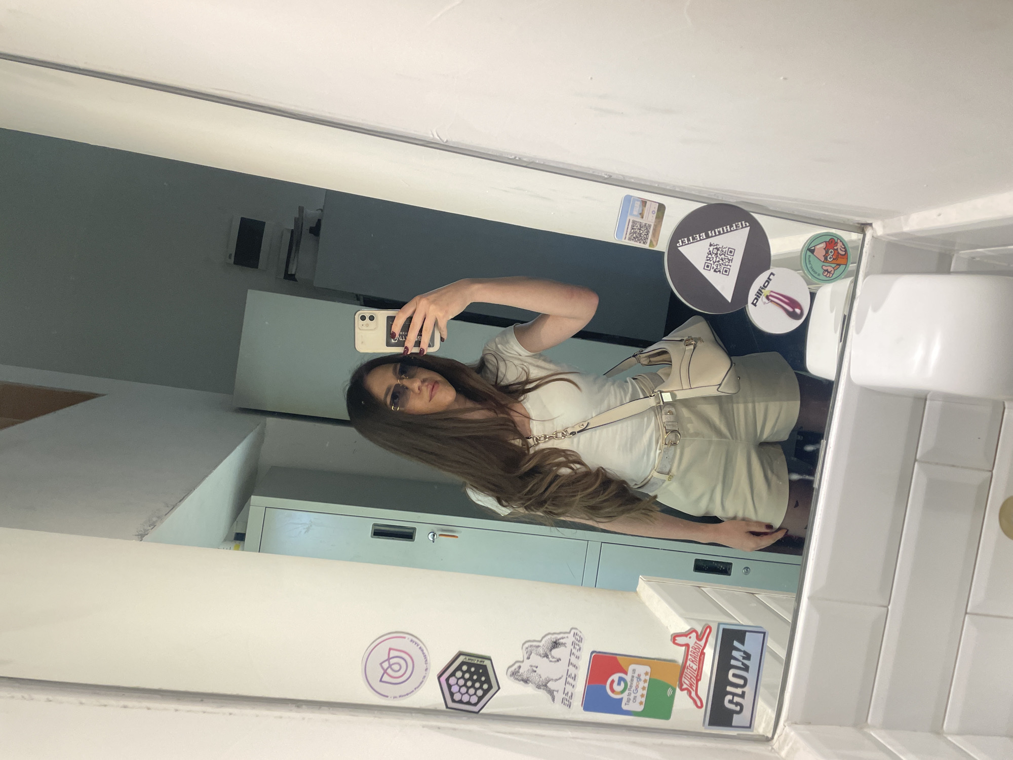 A person with long brown hair wearing sunglasses, a light-colored top, and shorts is taking a selfie in a mirror. The mirror is affixed to a wall with various stickers on its surface. The reflection shows a room with filing cabinets. The stickers include recognizable brand names like "WHITE RABBIT," "GLOW," and a Google review sticker. There is also a sticker with Cyrillic text ("ЧЁРНЫЙ ВЕТЕР") and the word "pillion". The location is likely a commercial space in Sofia, Bulgaria, considering the stickers and the language used.