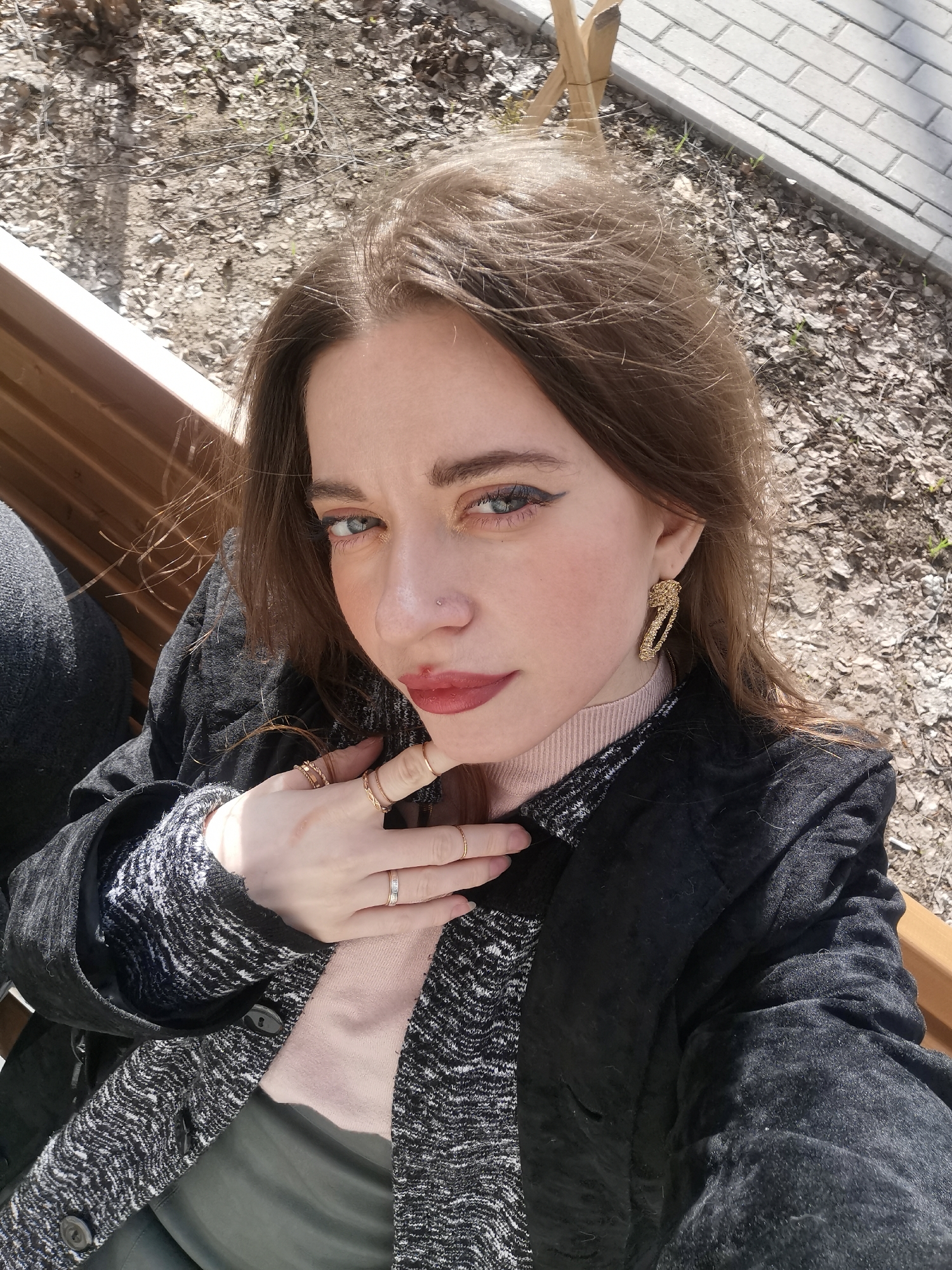 A close-up, eye-level selfie captures a young woman sitting outdoors on a sunny day. She has long, light brown hair, striking blue eyes with dramatic winged eyeliner, and reddish-brown lipstick. A small nose piercing is visible on her right nostril. She wears large, intricate gold-toned earrings and multiple gold-toned rings on her fingers, some with a twisted design.
She is dressed in a light pink turtleneck under a black and white patterned knit sweater, partly covered by a black blazer or jacket. Her right hand is raised to her chin and neck, with her ring-adorned fingers prominent.
The background suggests a park or an outdoor public area, with a wooden bench visible to the left. The ground is covered with dried leaves, twigs, and sparse patches of grass. In the upper right, a section of light grey paved bricks is visible, alongside a wooden post. The strong highlights and shadows indicate bright daylight. Her expression is direct and intense, looking straight at the camera. No text is visible in the image.