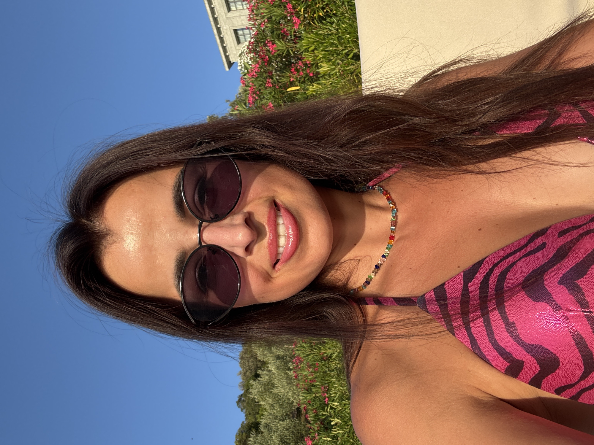 A woman is taking a selfie outdoors on a sunny day. She has long, dark brown hair and is wearing sunglasses and a pink bikini top with a zebra print. She is also wearing a colorful beaded necklace. In the background, there is a wall, a lush green hillside with trees and pink flowers, and a building with a light-colored facade and dark windows. The sky is clear and blue.