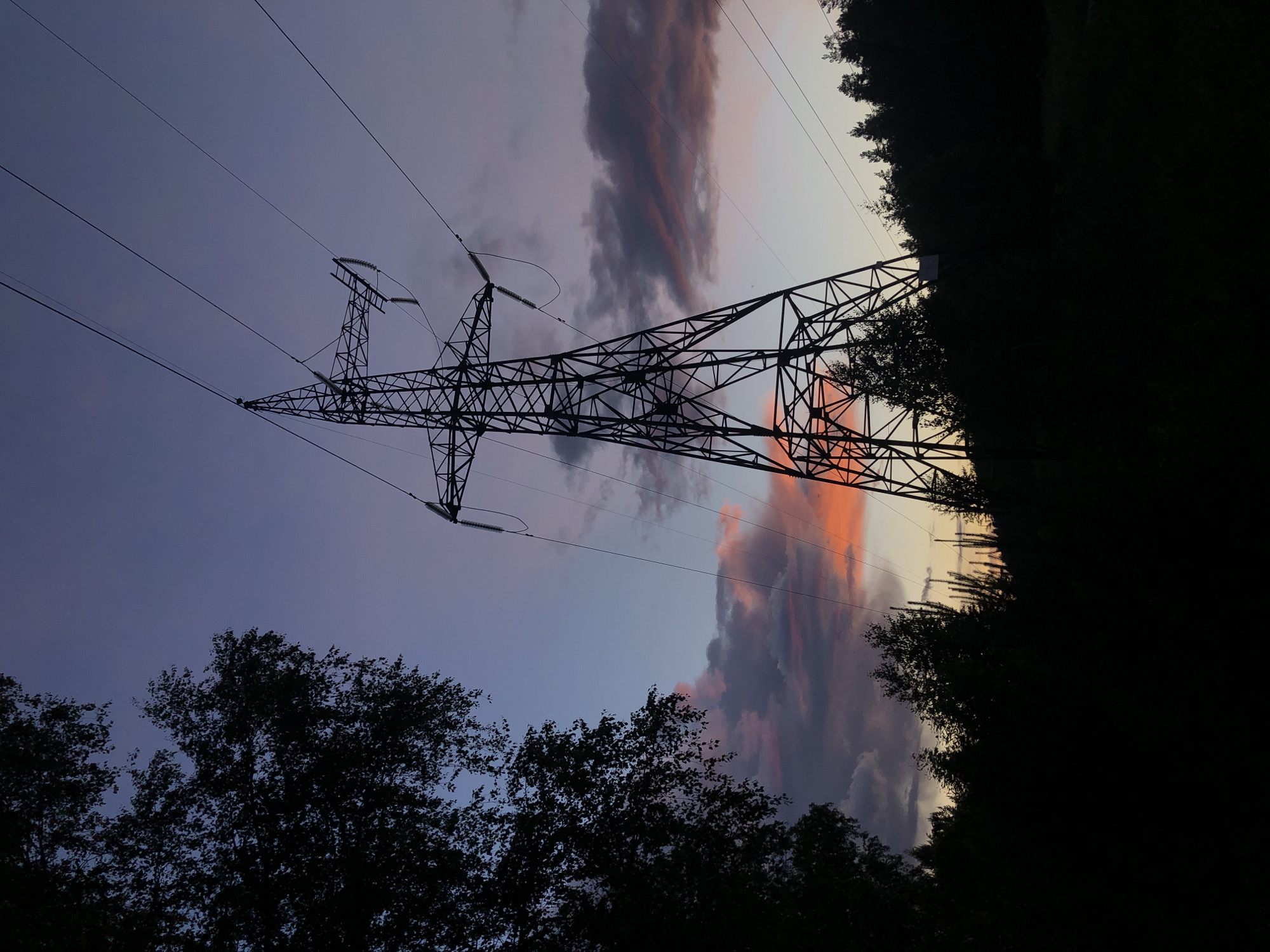 A low-angle shot depicts a transmission tower silhouetted against a twilight sky. The sky exhibits hues of purple, pink, and orange, indicating sunset or sunrise. The tower's latticework structure is complex and intricate, with power lines stretching outward. Silhouetted trees are visible along the bottom and right edges of the frame. The location is Noviy, Russia.