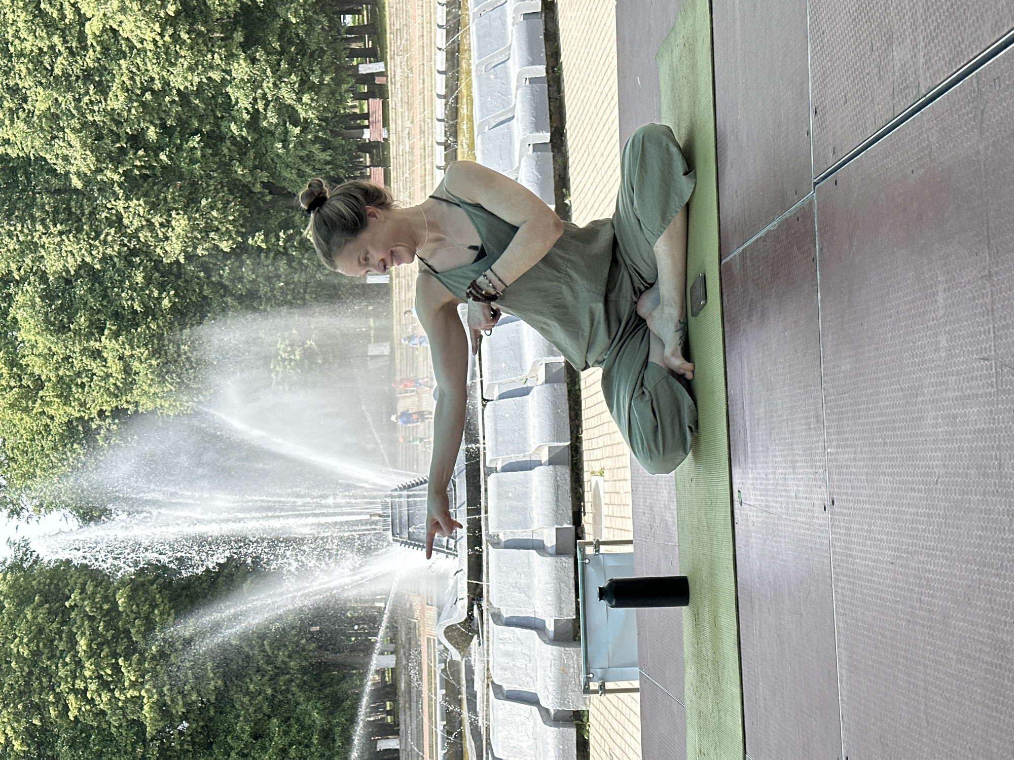 A woman in a green jumpsuit is sitting cross-legged on a green yoga mat on a stage. She is pointing to the left with her right hand, her gaze directed in the same direction. Her left arm is bent, with her hand near her chest. She has a black object, possibly a water bottle, to her left. Behind her, a large fountain sprays water high into the air, with lush green trees and benches visible in the background. The scene appears to be an outdoor setting in a park, potentially the Theater Square in Vladimir, Russia, known for its fountains and green spaces. The woman's posture suggests she might be engaged in a yoga demonstration or lesson.