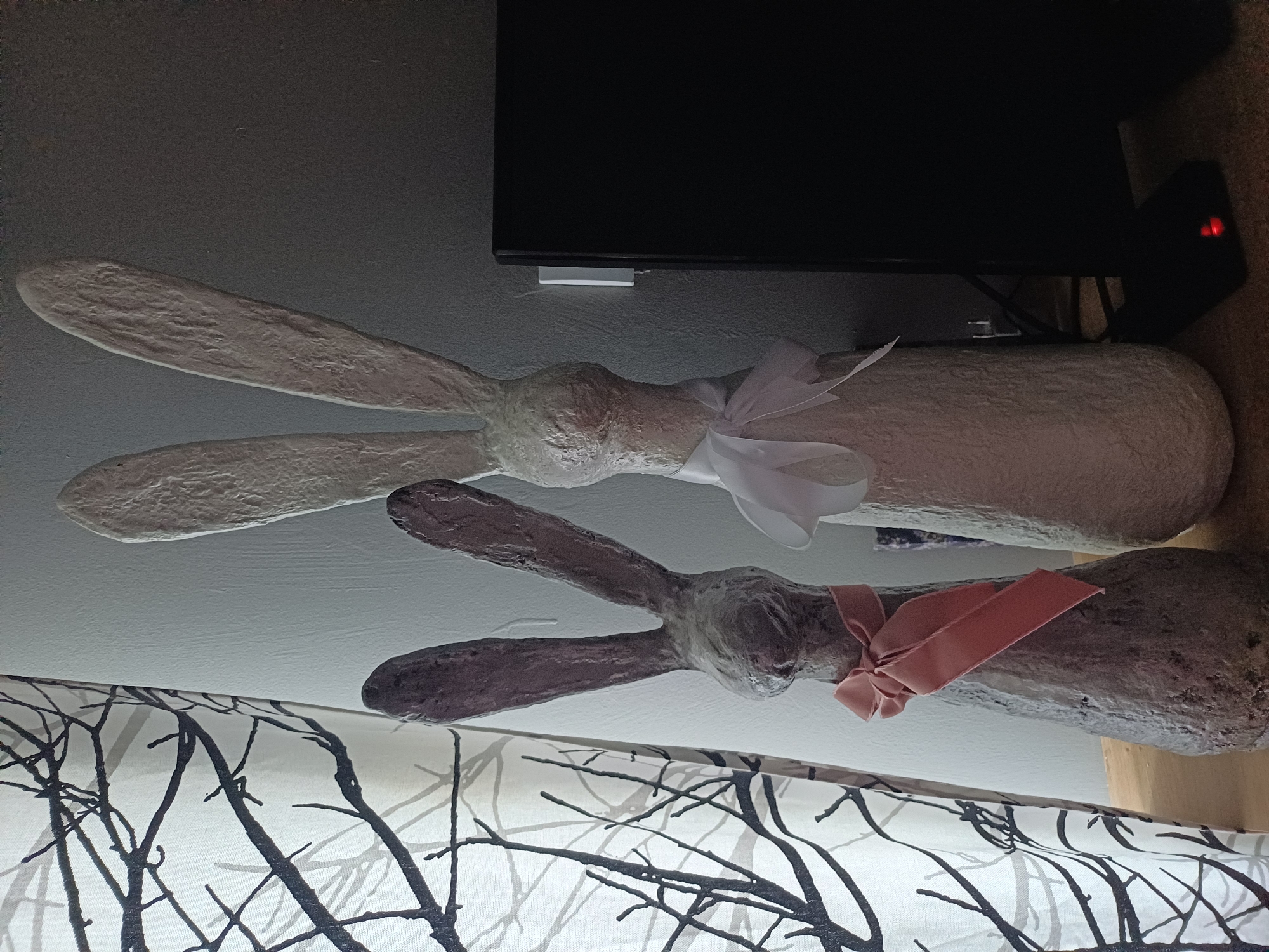 In Sulymivka, Ukraine, two Easter bunny decorations stand on a wooden surface. The bunny on the left is dark gray with a pink ribbon tied around its neck. The bunny on the right is light cream-colored with a white ribbon around its neck. To the left of the bunnies is a sheer curtain with a black tree branch pattern. To the right of the bunnies is a black electronic device. The wall behind the bunnies is a textured gray. The scene is lit from the left, casting shadows to the right.